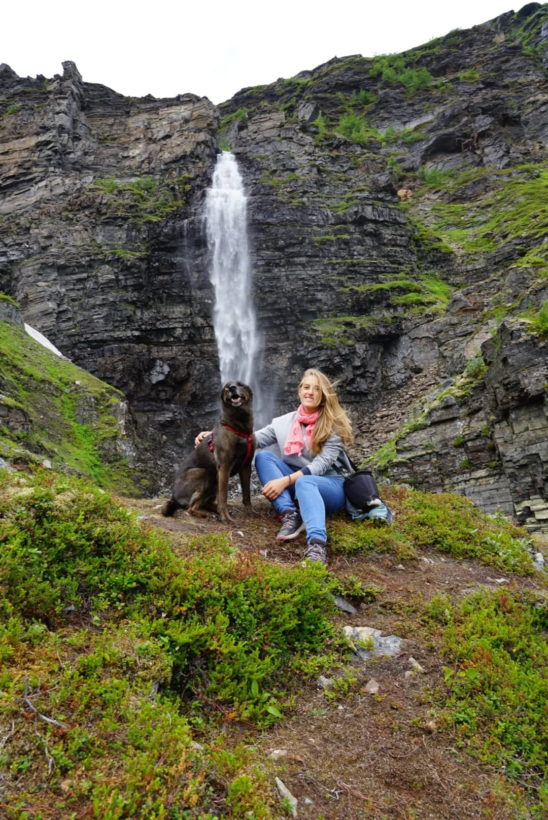 Mujer y perro posando frente a una cascada en zona de montaña con vegetación y rocas.