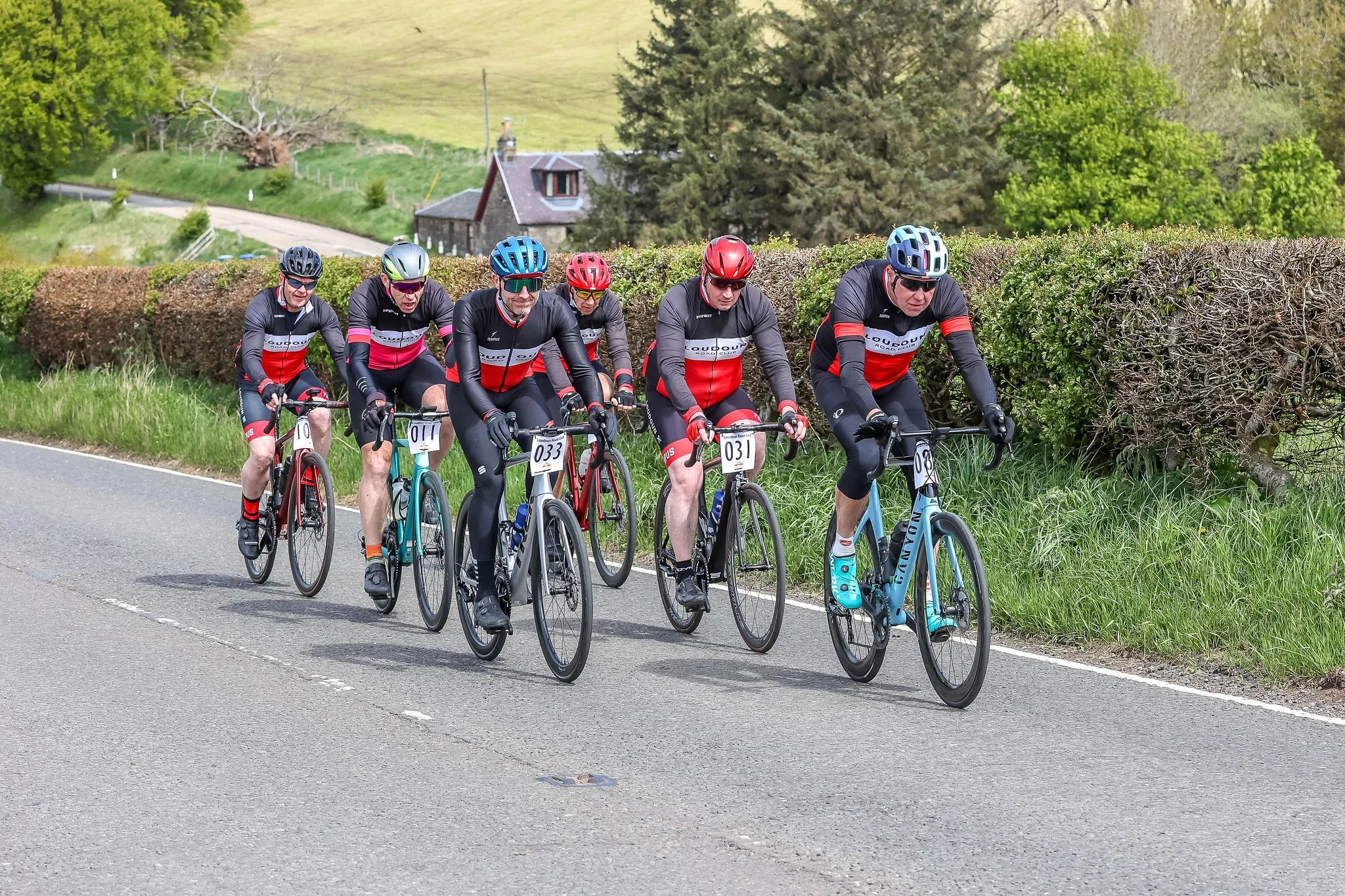 Cycling group riding on a rural road with trees and houses in the background.