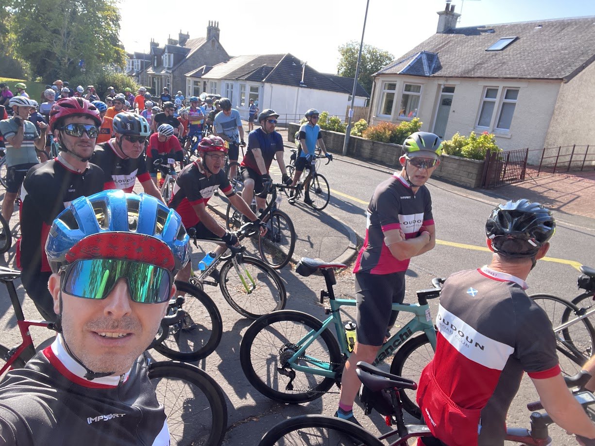 Group of cyclists gathered outdoors, some wearing helmets and sunglasses, standing with their bikes in a sunny residential area with houses and greenery in the background.