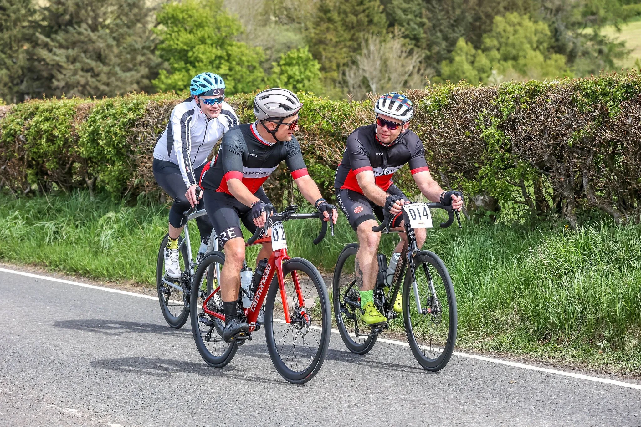 Three cyclists riding bikes along a scenic rural road, surrounded by green trees and bushes.