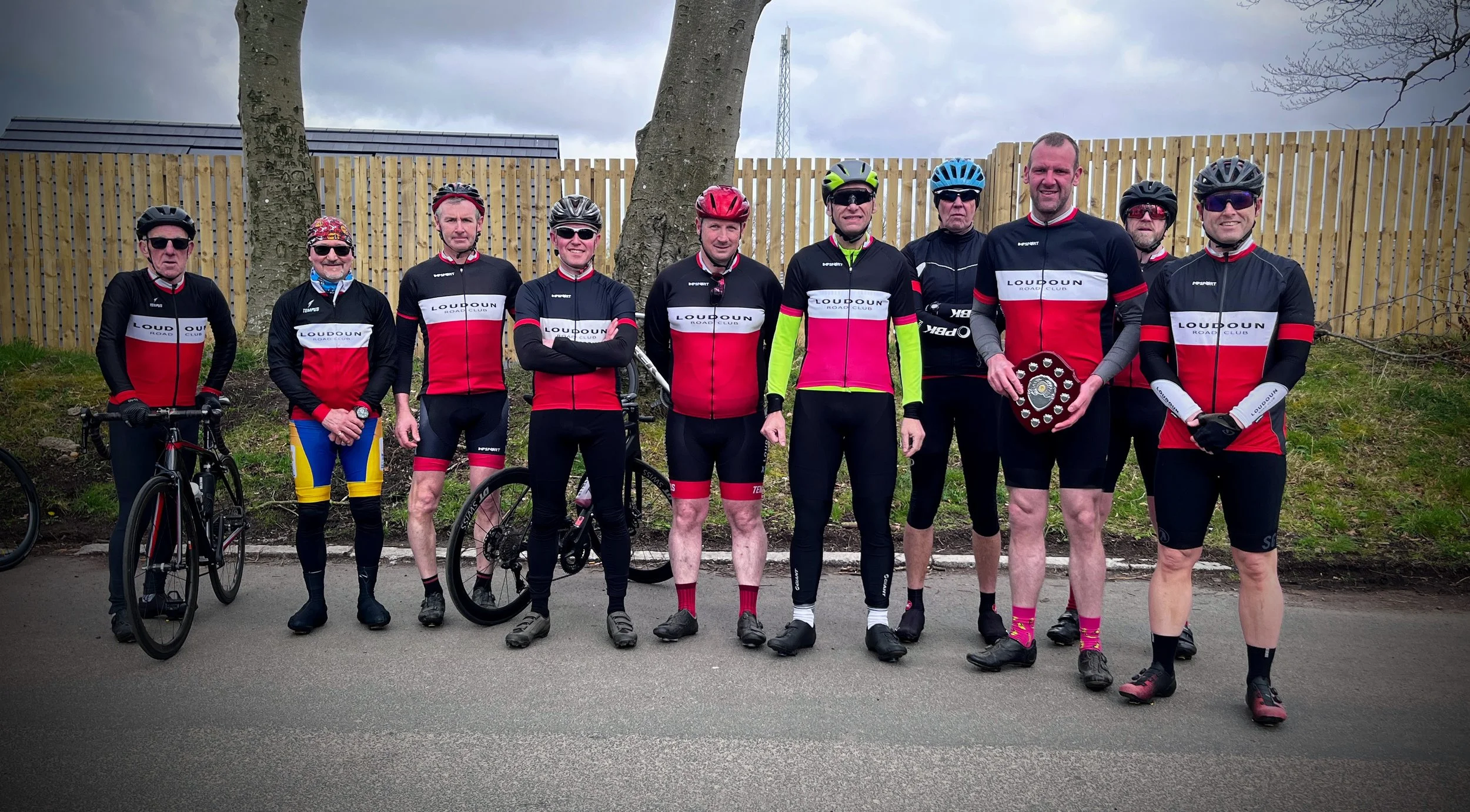 A group of eleven adult cyclists in matching team uniforms posing outdoors on a paved surface, with a wooden fence and trees behind them, some holding bicycles, one holding a shield-shaped trophy.