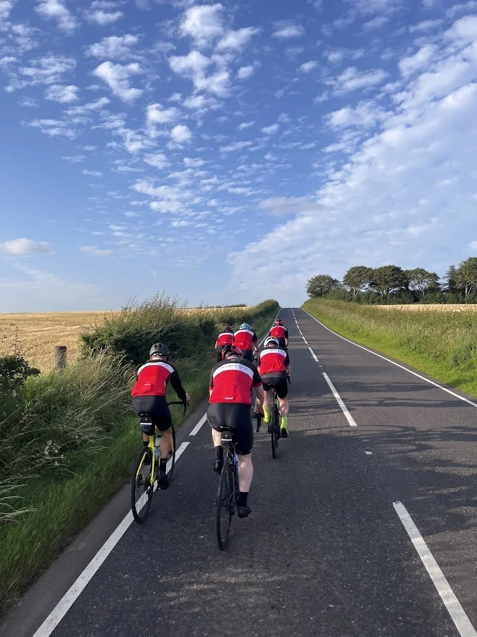 A group of cyclists riding on a rural road under a blue sky with scattered clouds.