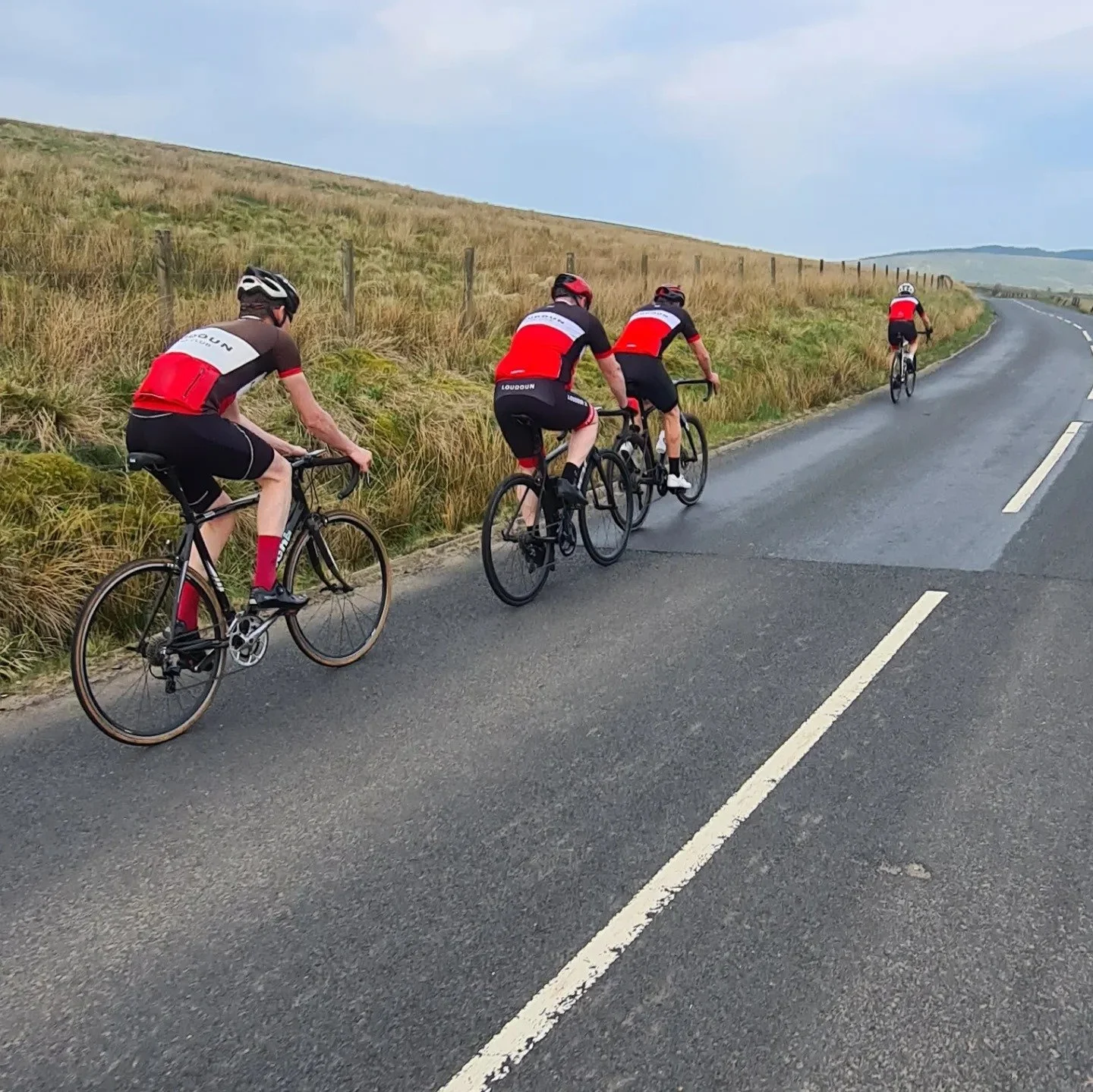 Four cyclists riding on a paved country road, surrounded by grassy hills and a fence, with cloudy skies overhead.