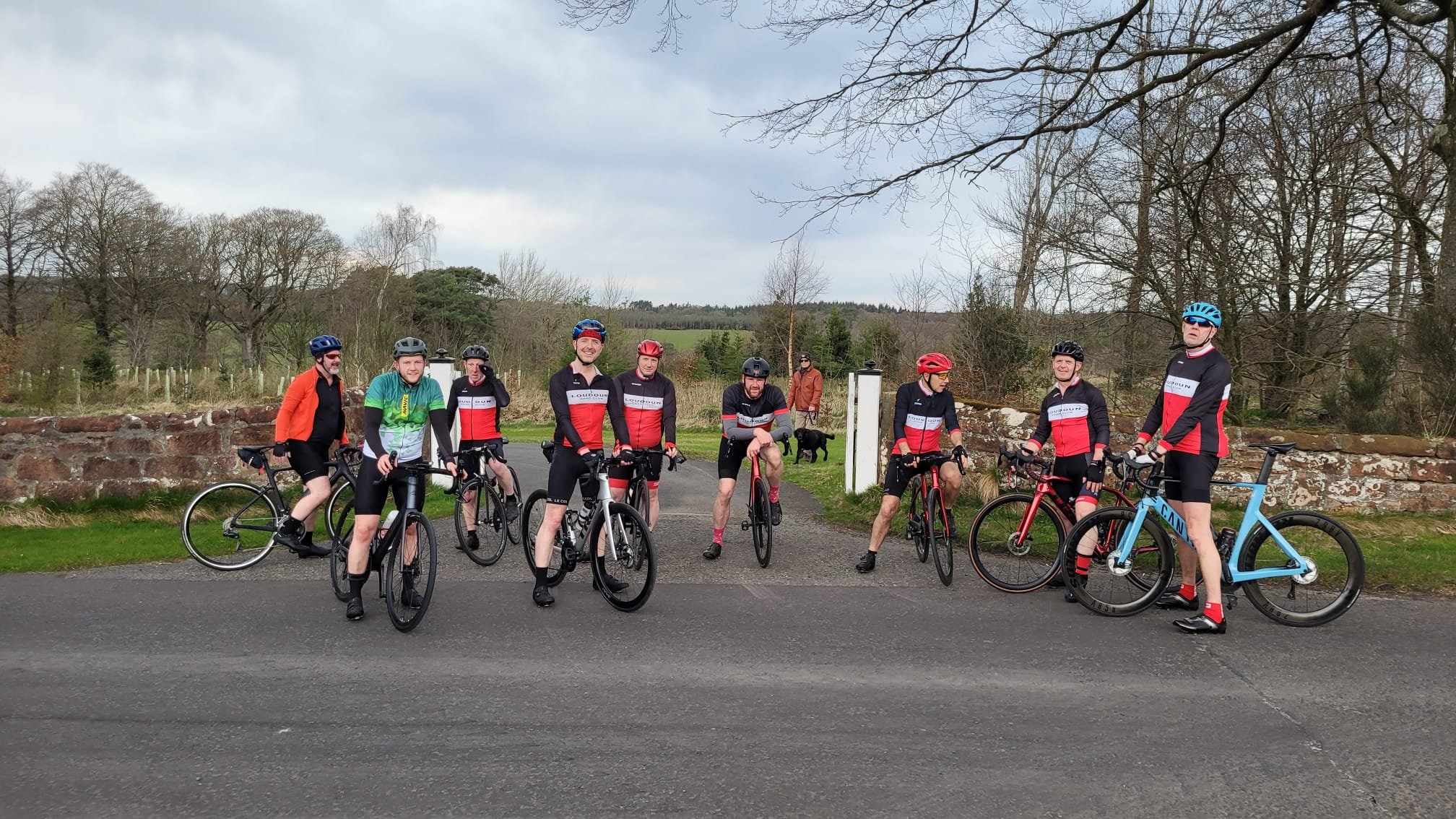 A group of nine cyclists, six men and three women, standing on the side of a road with their bikes. They are dressed in cycling gear with helmets and sunglasses, and are smiling or looking relaxed. The background features a rural landscape with leafl