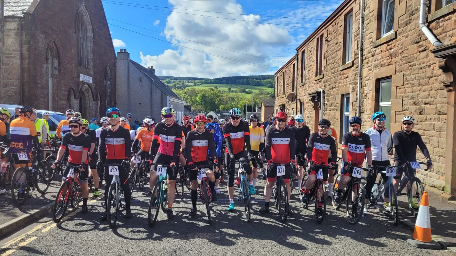 Group of cyclists in race gear with helmets, standing with bikes on a street in a small town with stone buildings and green hills in the background, sunny day.