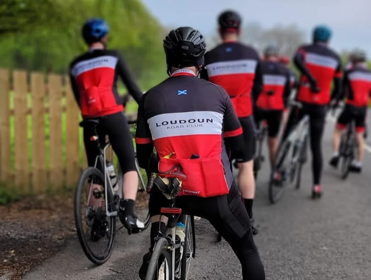 A group of cyclists from the Loudoun Road Club resting on a paved path, wearing black and red cycling jerseys with the club's name, helmets, and cycling gear.