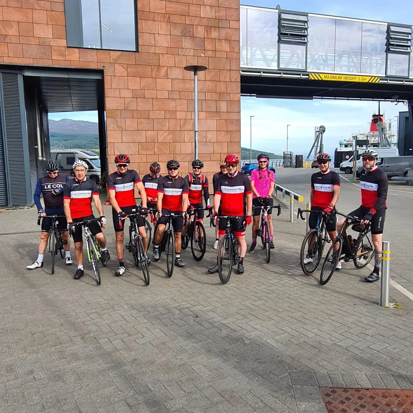 Group of ten cyclists in matching black and red cycling jerseys and helmets, with some also wearing sunglasses, standing with bicycles outside building near port with ships and mountains in background.
