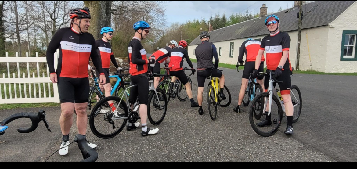 A group of cyclists in matching team jerseys and helmets gathered on a rural road, standing with their bikes, some talking and one preparing to ride.