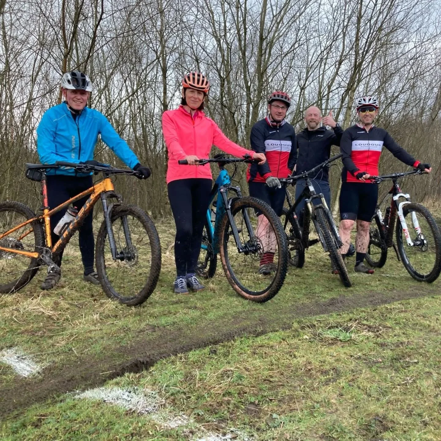 Five people standing with mountain bikes outdoors on a muddy trail, wearing helmets, cycling gear, and rainy weather clothing, in a leafless wooded area.