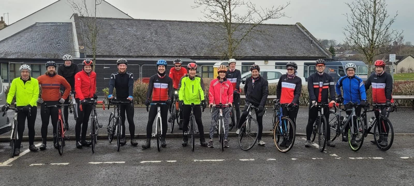 Group of fifteen cyclists standing with their bikes in a parking lot