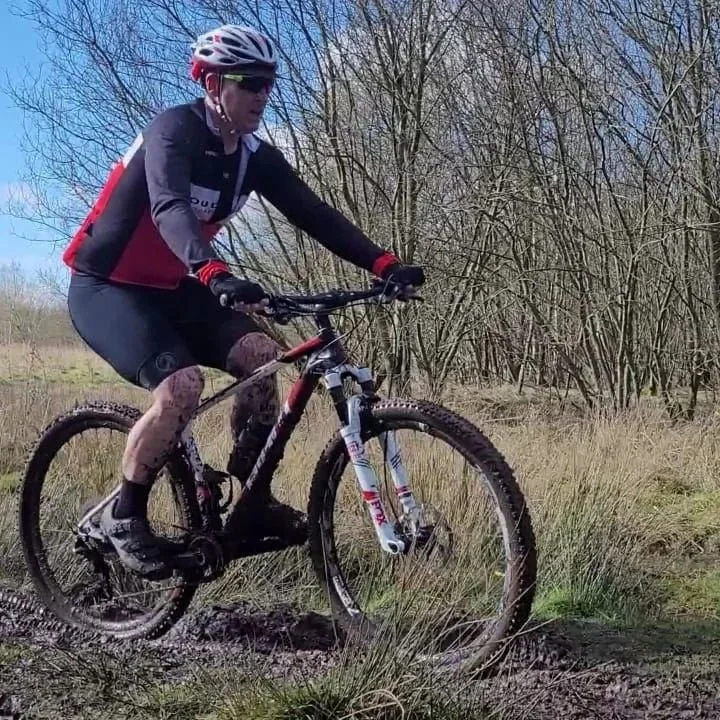 A person riding a mountain bike on a dirt trail in a wooded area, wearing a helmet, sunglasses, and cycling gear.
