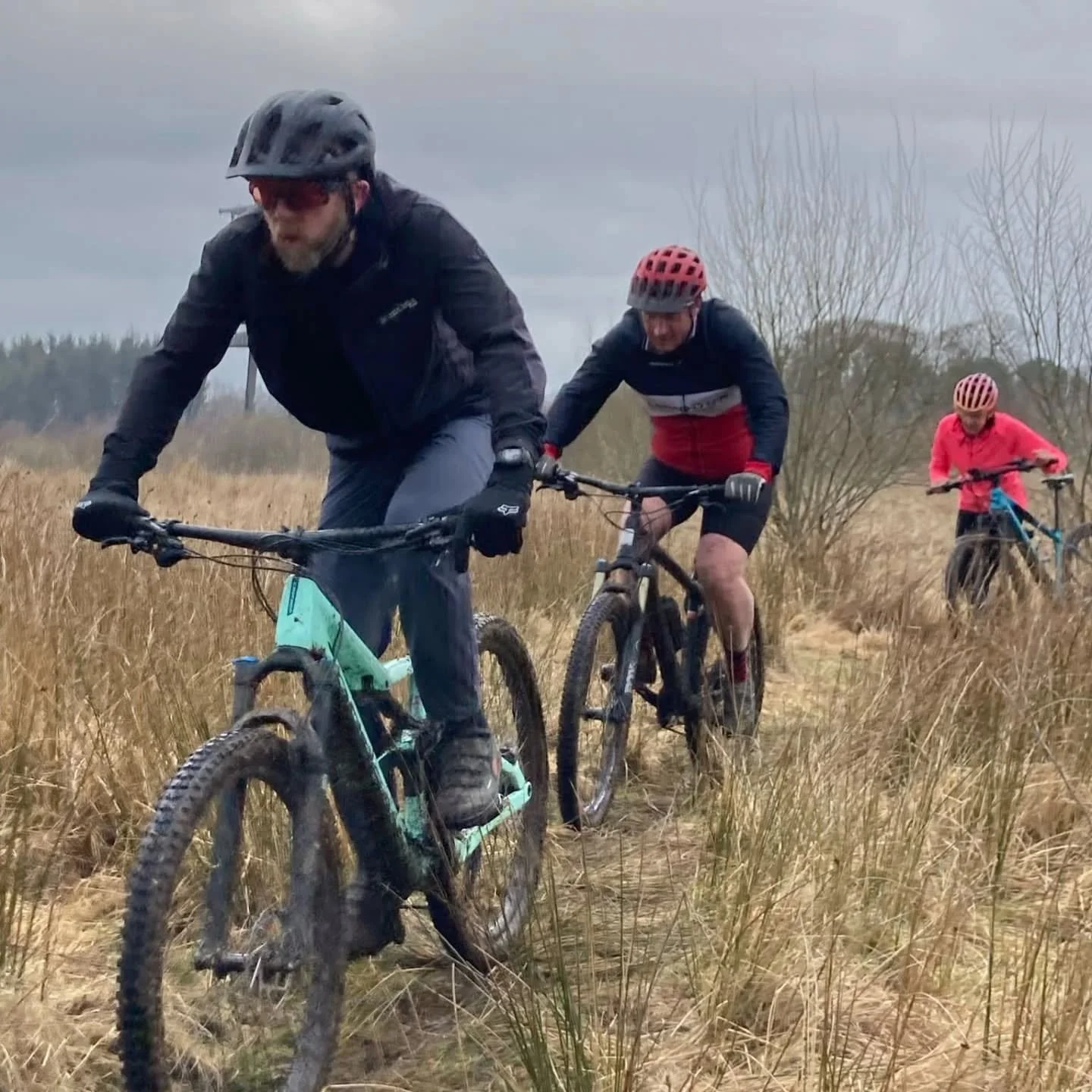 Three people mountain biking through a grassy field on a cloudy day, each wearing helmets and outdoor gear.