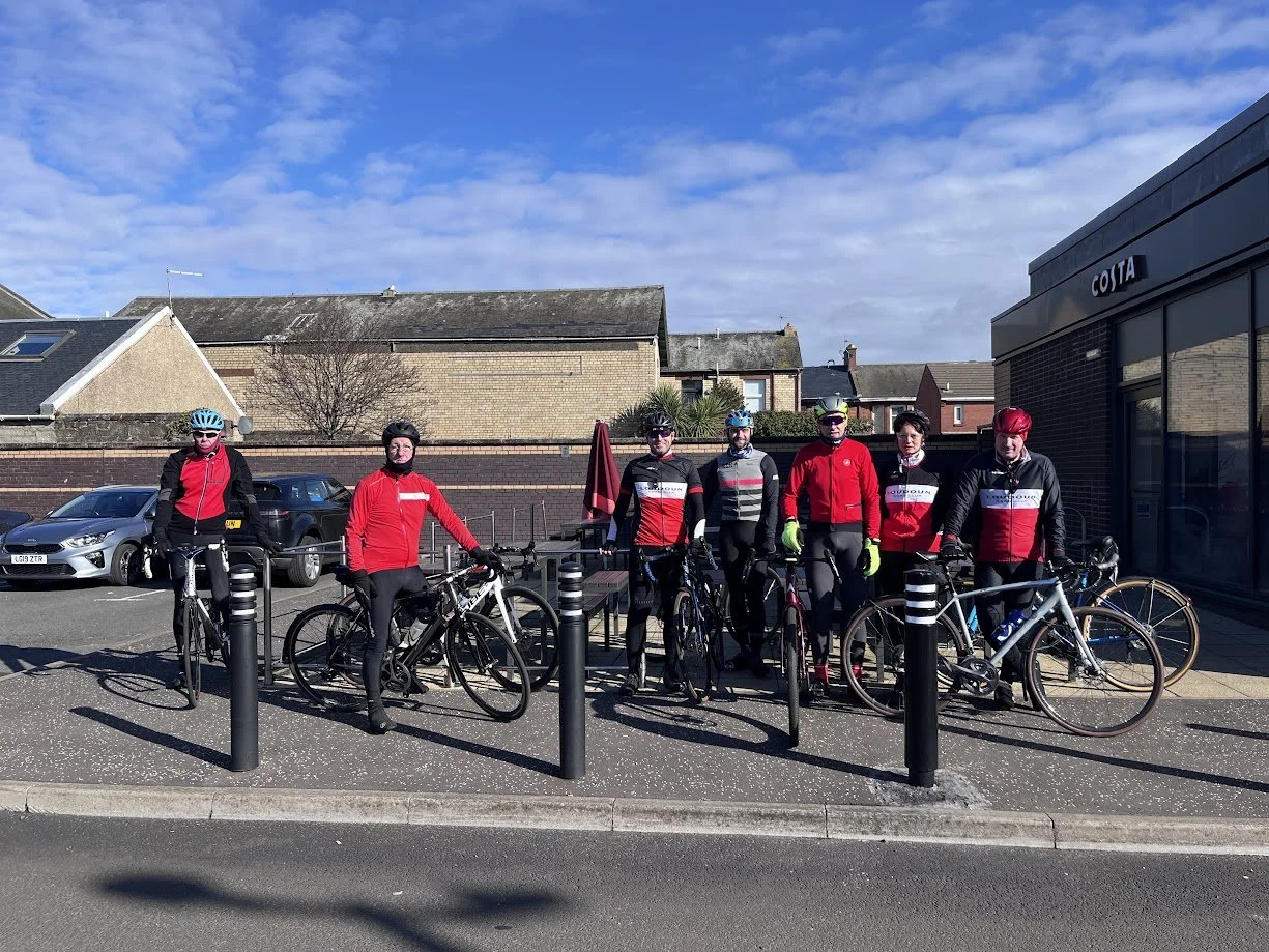 Group of seven cyclists standing with their bikes outside a cafe on a sunny day, wearing cycling gear and helmets.