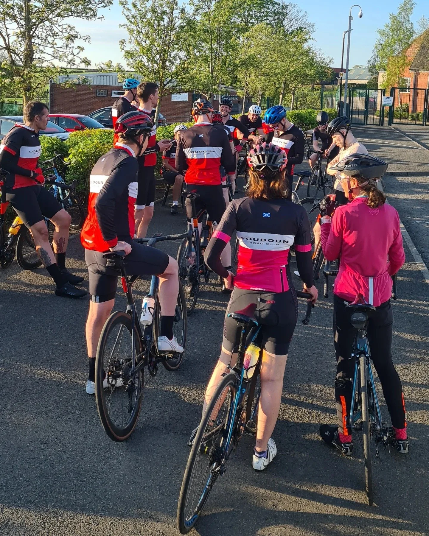 Group of cyclists wearing matching black, red, and white cycling outfits with the logo 'LOUDOUN ROAD CLUB,' gathered outside on a sunny day, some standing and some sitting on bikes, with trees and parked cars in the background.