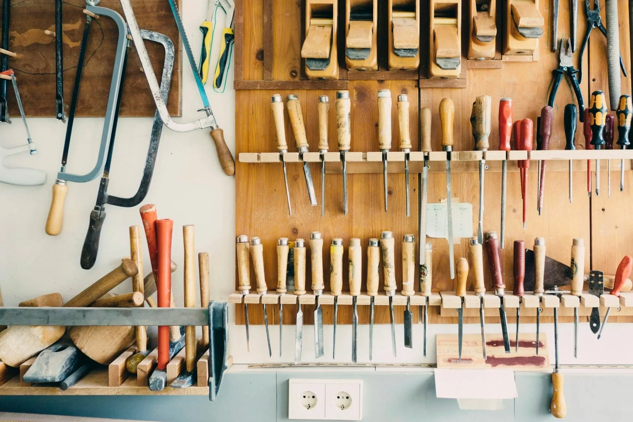 A wall-mounted organized collection of woodworking tools including hand saws, chisels, screwdrivers, files, and a hacksaw, with some tools hanging on a wooden pegboard and others placed on a shelf below, with power outlets visible at the bottom.