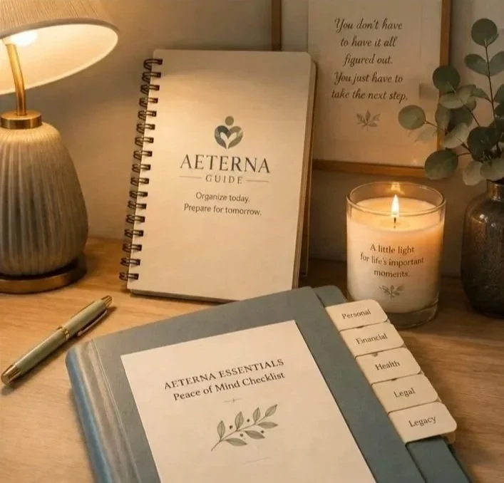 A desk setup with a spiral notebook titled 'Aeterna Guide', a pen, a blue folder labeled 'Aeterna Essentials Peace of Mind Checklist' with labeled tabs for 'Personal,' 'Financial,' 'Health,' 'Legal,' and 'Legacy'; a lit candle with a quote, a framed inspirational quote, a table lamp, and a vase with greenery.