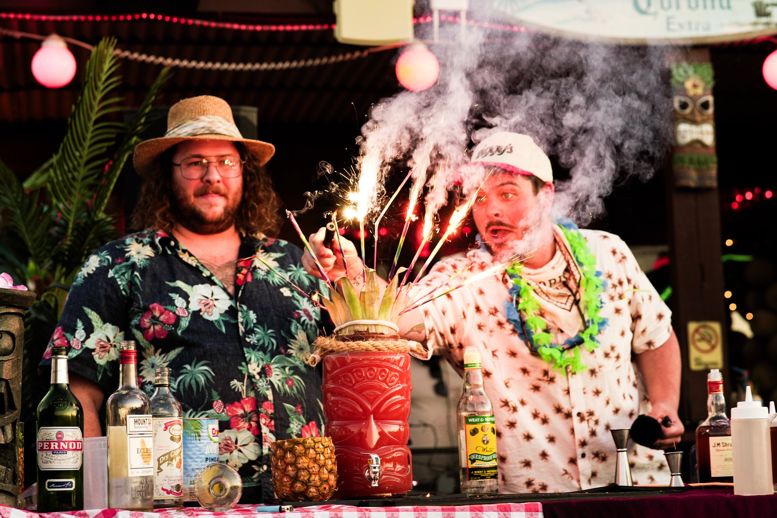 Two men in Hawaiian shirts and hats celebrating with sparklers and smoke at a festive event with liquor bottles and tropical decorations.