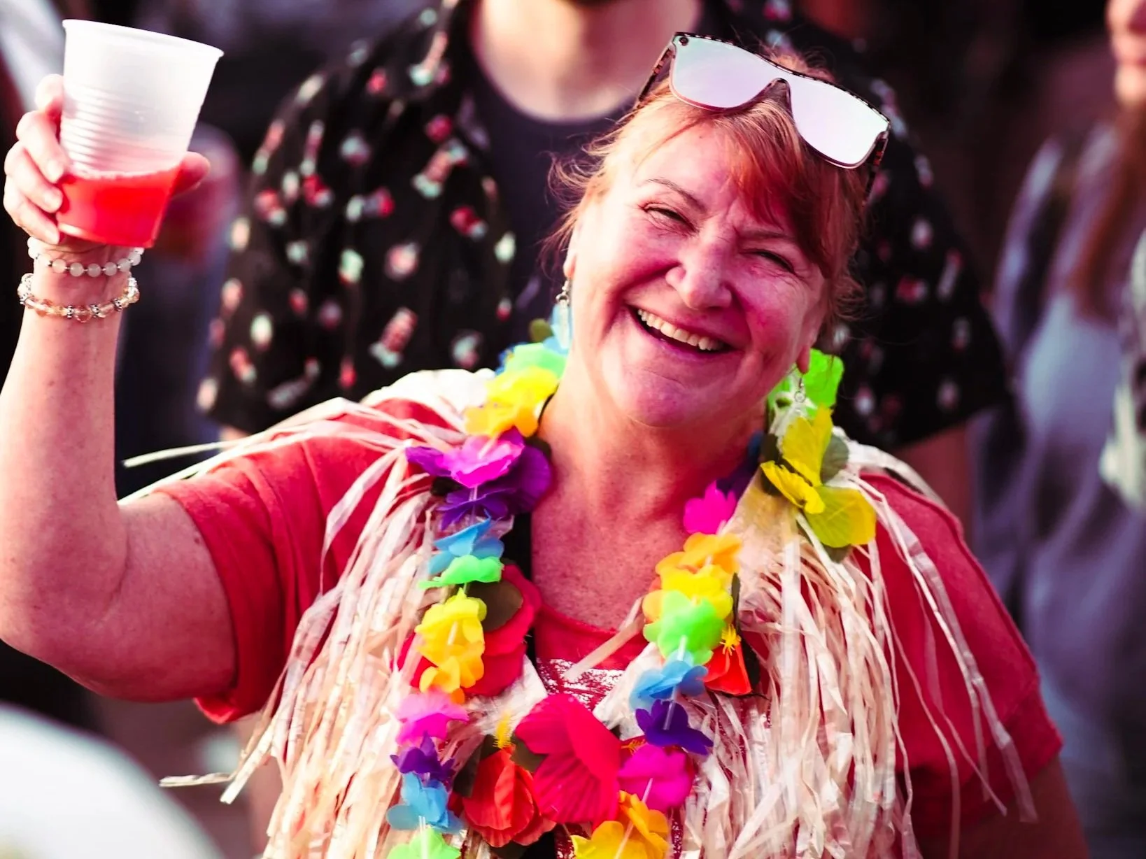 A woman smiling and holding a red drink in a plastic cup, wearing colorful leis and sunglasses on her head at a party or celebration.