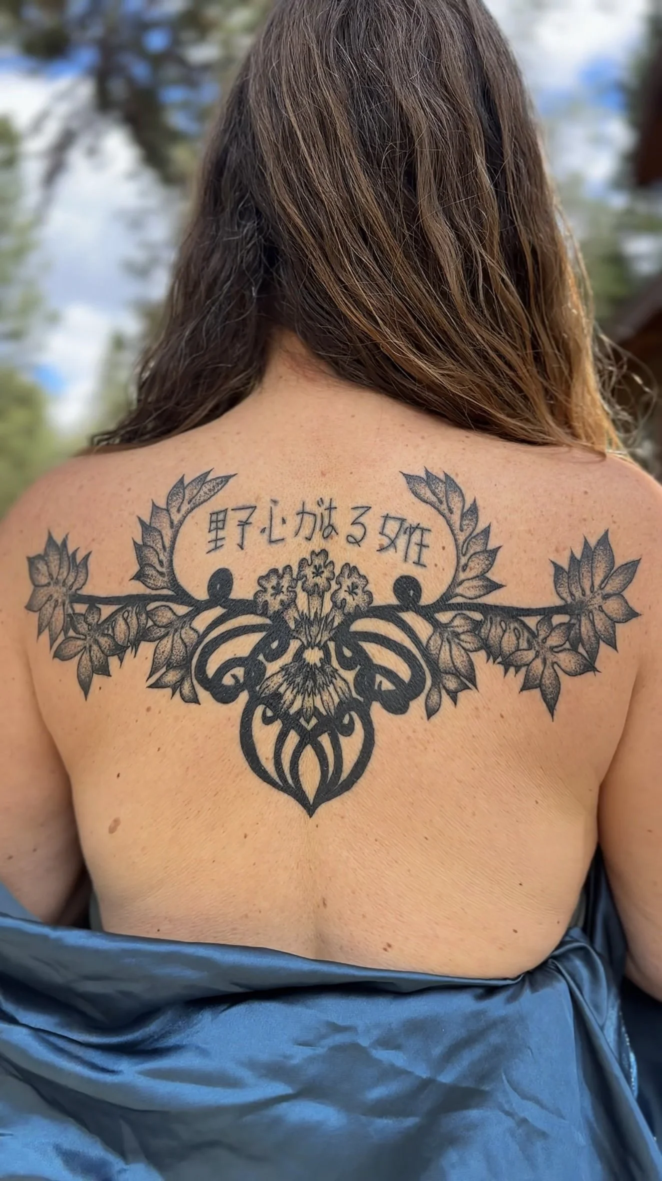 Back of a woman with brown hair, showing a large, intricate black tattoo on her upper back. The tattoo features a central symmetrical design surrounded by extending floral and leaf motifs and contains Japanese characters.