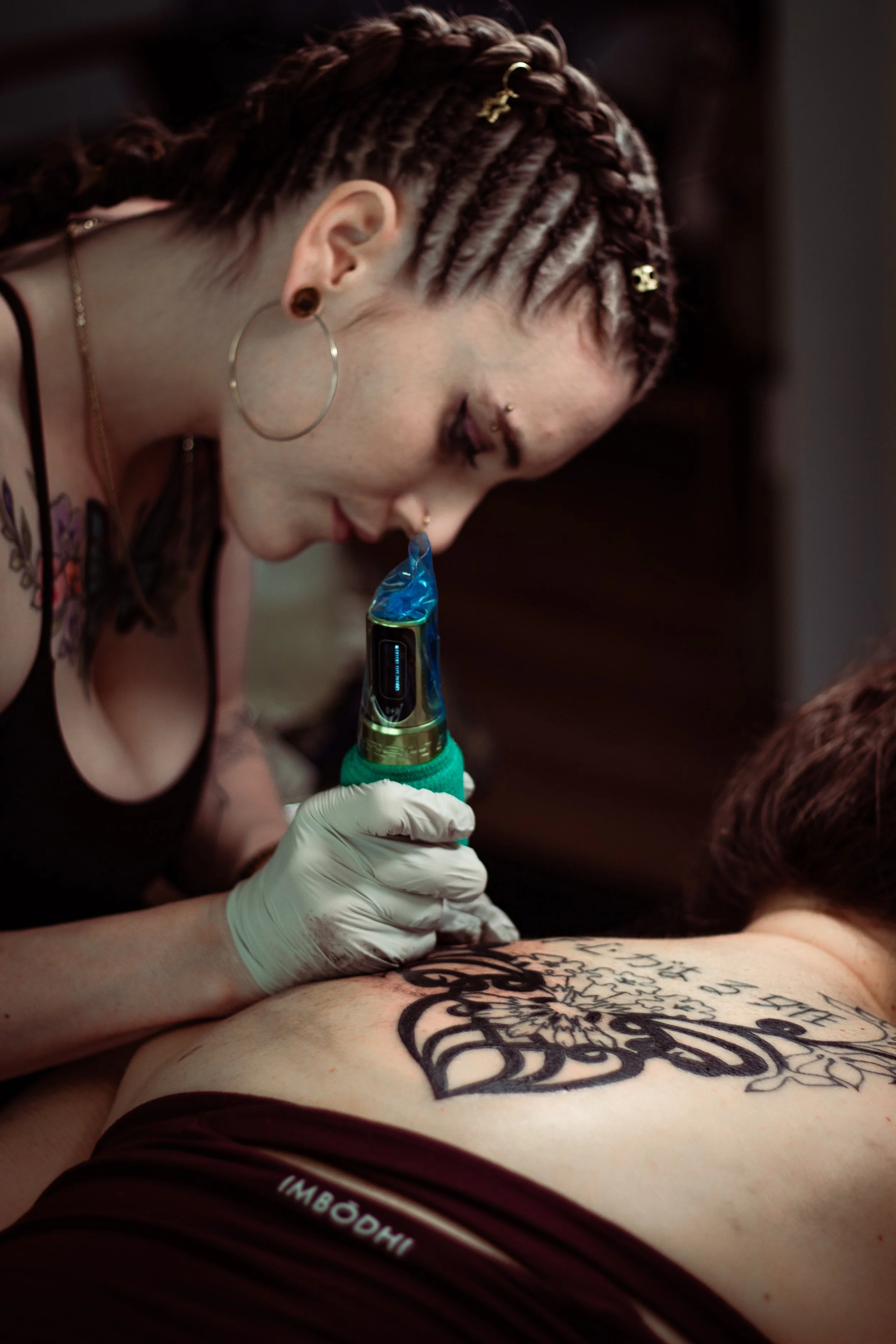 A tattoo artist with multiple nose rings and large hoop earrings is tattooing a person's stomach, which has a large, intricate black tattoo. The person wearing maroon shorts is lying face down, and the tattoo artist is using a tattoo machine while we
