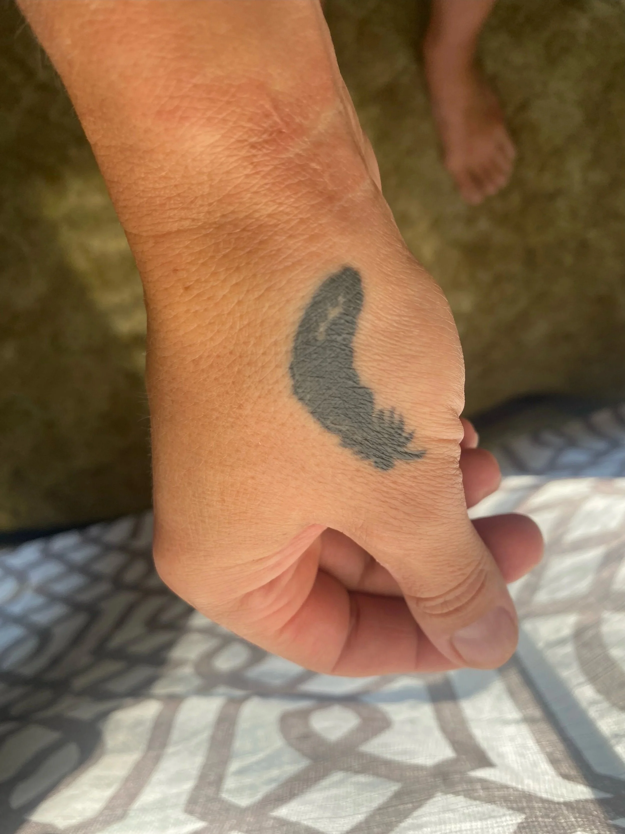 Close-up of a hand with a black tattoo on the wrist, in front of a patterned fabric background.