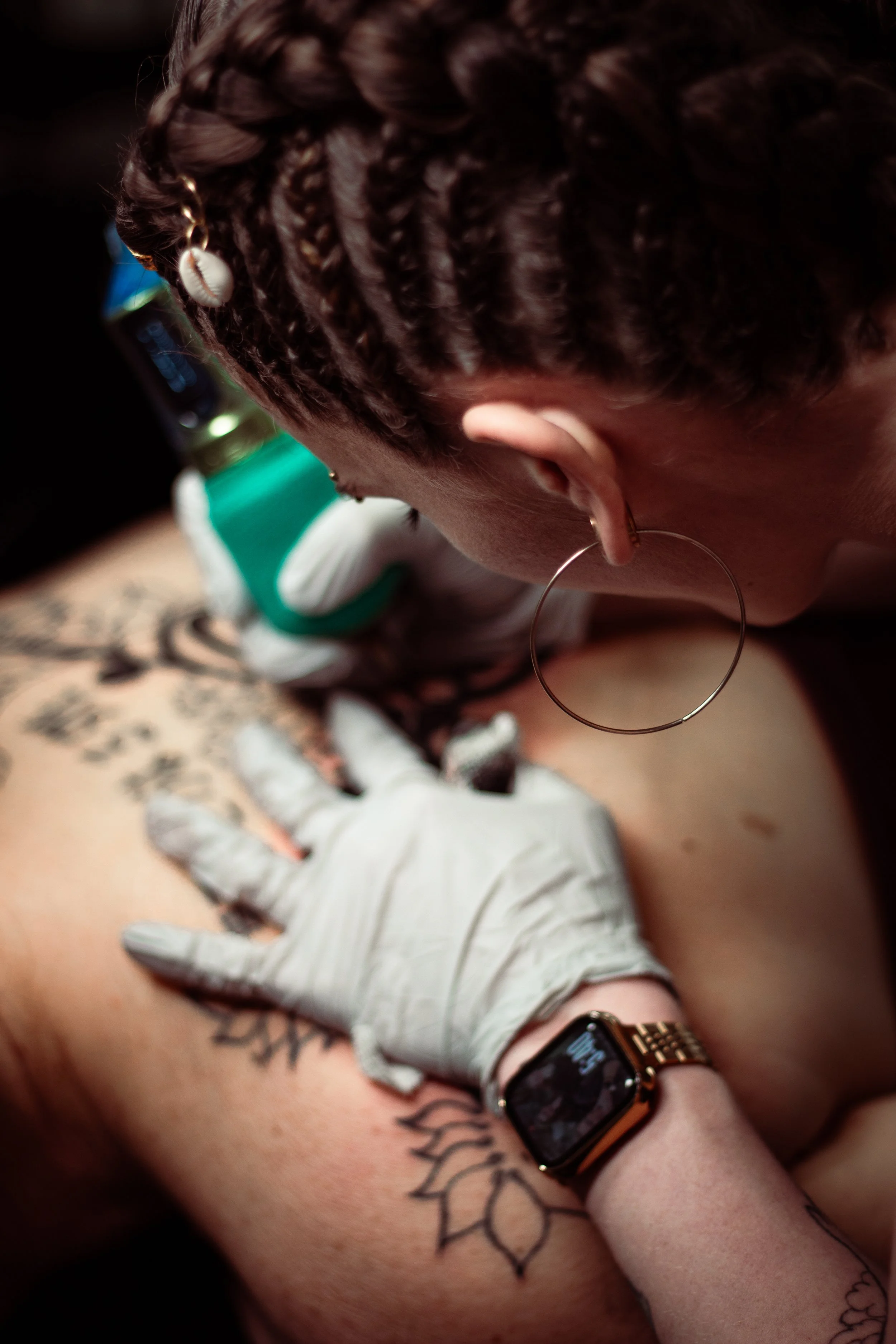 Close-up of a person's head with braided hair, wearing a hoop earring, with a tattoo visible on their arm, and a smartwatch on their wrist. Someone's gloved hand is visible, possibly tending to the person's arm.