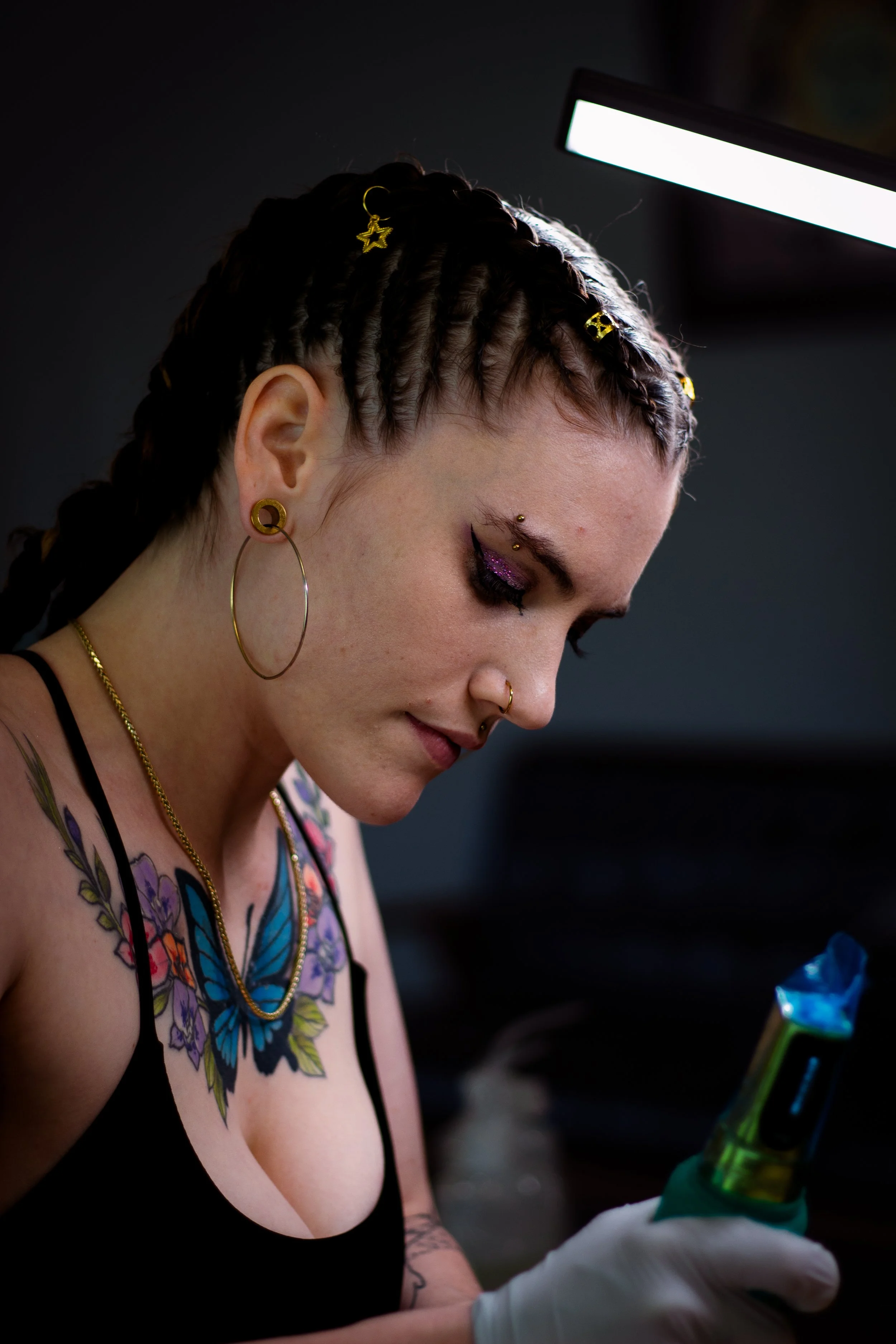 A woman with tattoos on her chest and arms, wearing large hoop earrings, a nose ring, and colorful jewelry, is looking at her phone while holding a tattoo machine in her gloved hand.