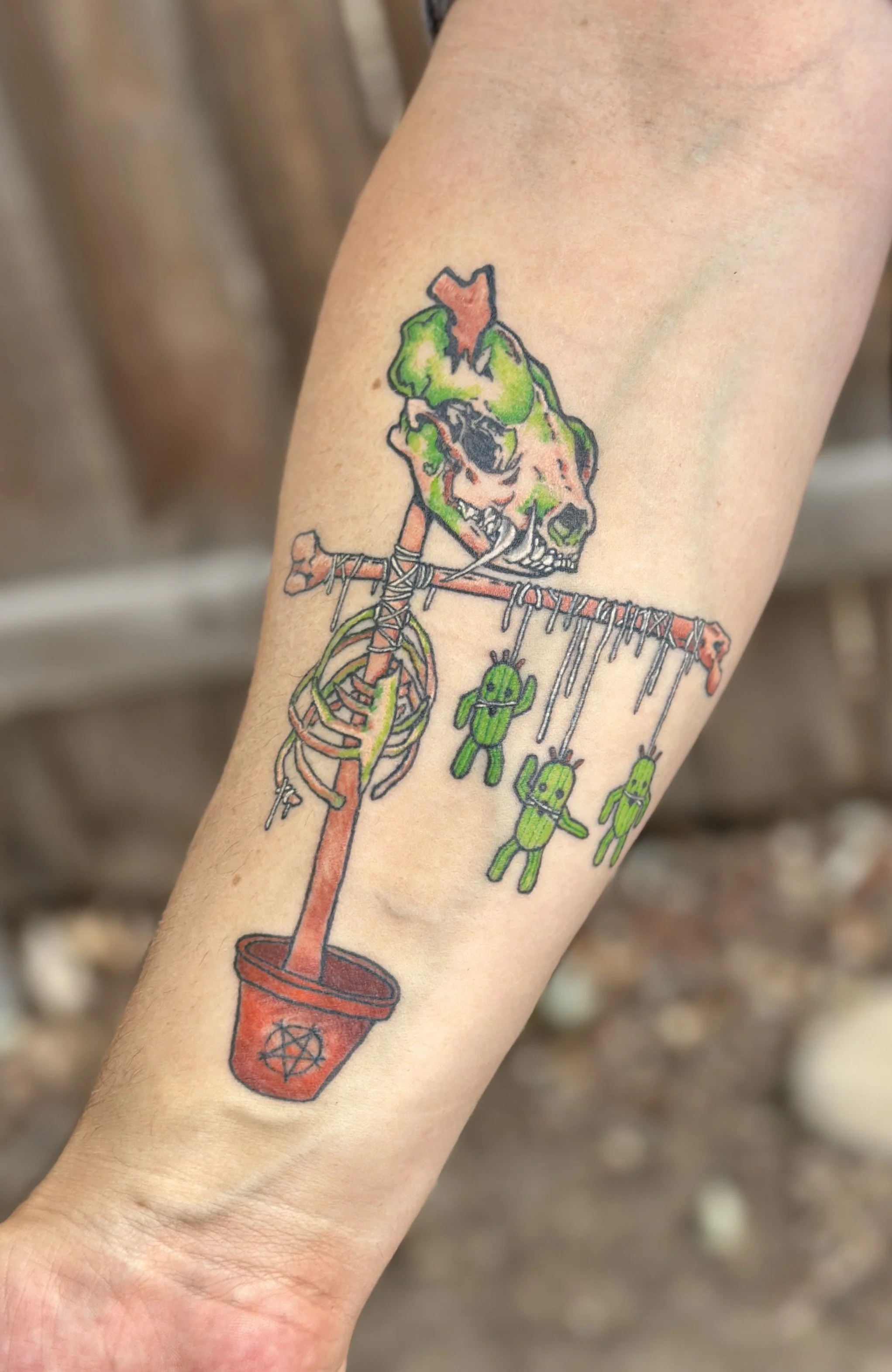 Tattoo of a skeleton with a green face holding a sword, standing over a red bucket with a pentagram symbol on it. The skeleton's basket has small green figures hanging from it.