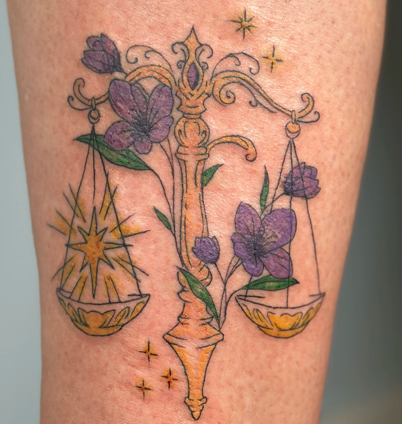Tattoo of a balanced scale with purple flowers, green leaves, and stars around it