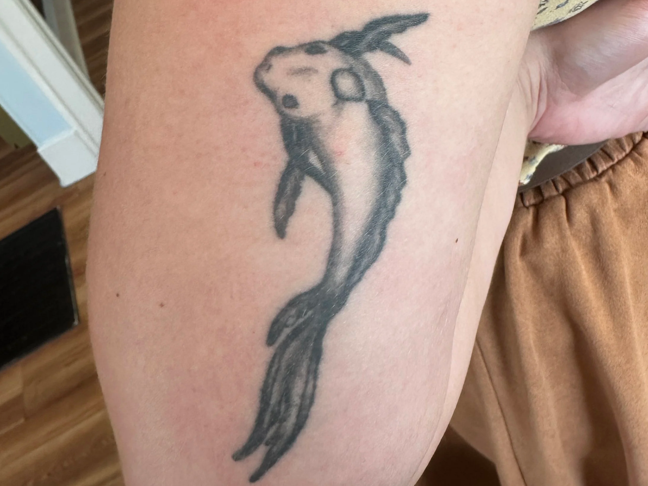 Close-up of a black and gray dragon tattoo on a person's arm, with a background of a wooden floor and part of a light-colored wall.