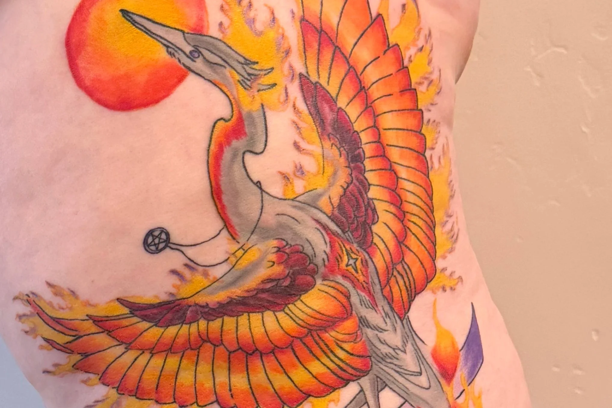 Colorful tattoo of a phoenix bird with orange and yellow flames on a person’s skin.