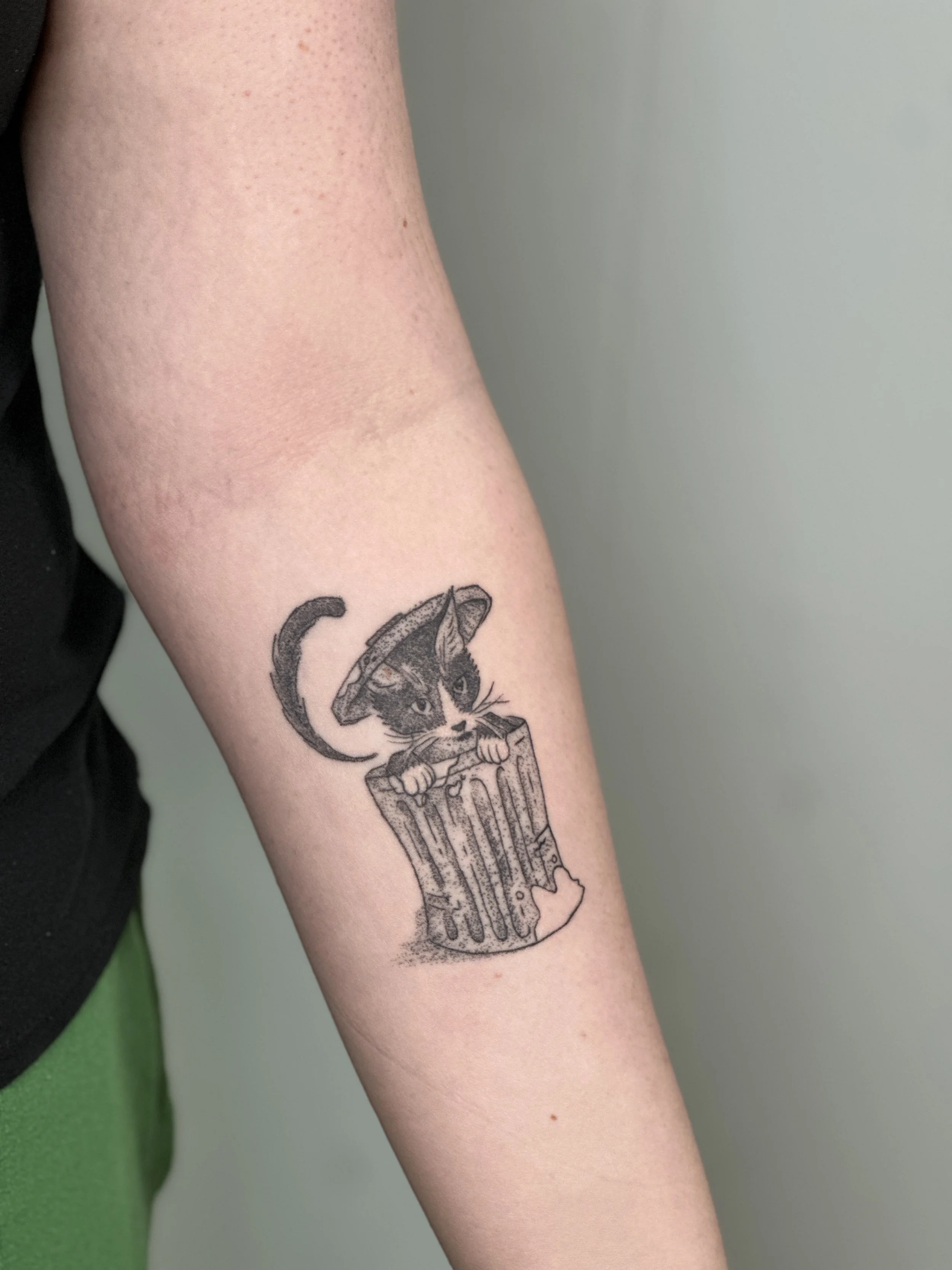 Tattoo of a cat with a hat in a trash can on a person's forearm.