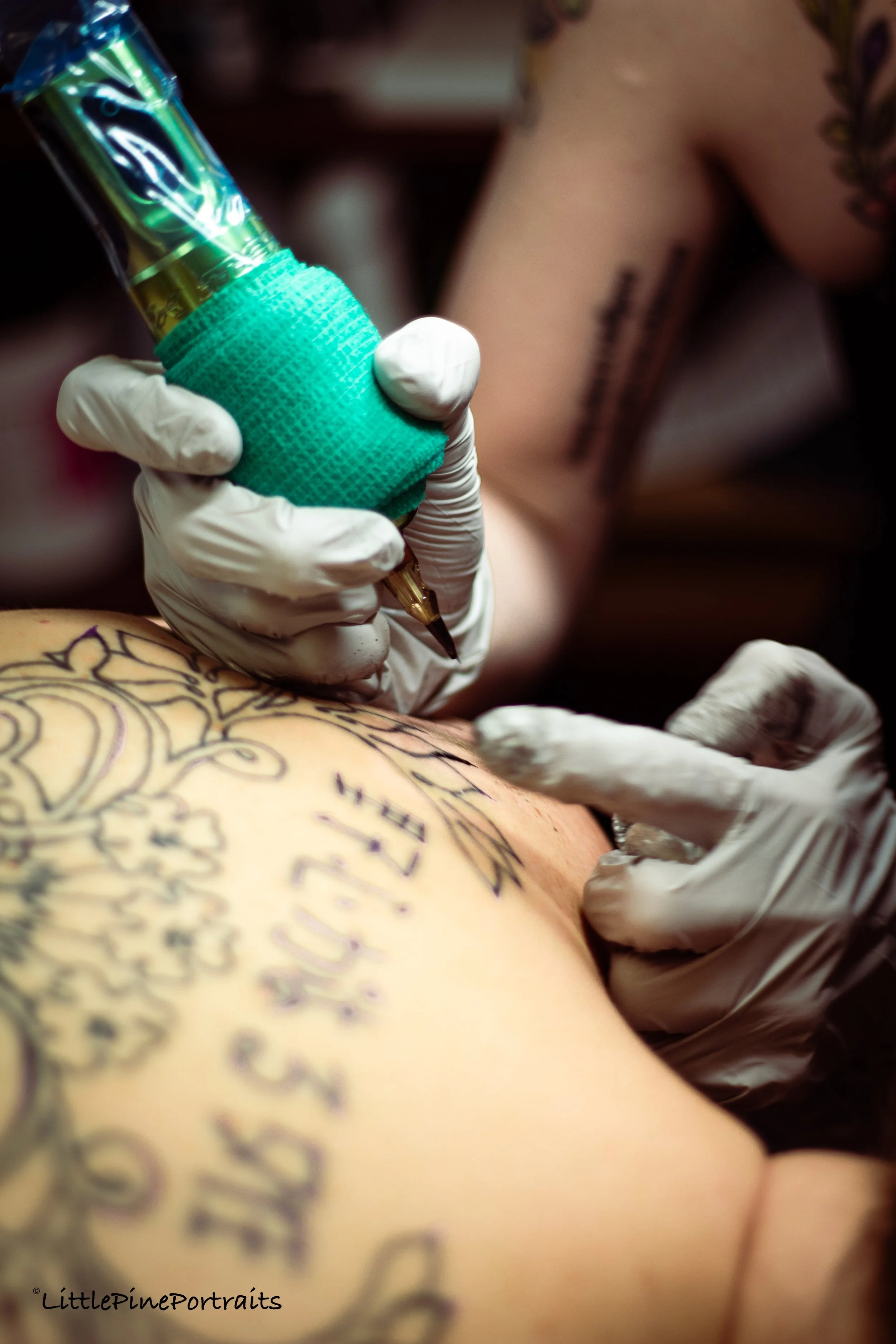 A tattoo artist, wearing white gloves, is tattooing a design onto a person's arm. The person has multiple tattoos, including a large, intricate script and decorative elements.