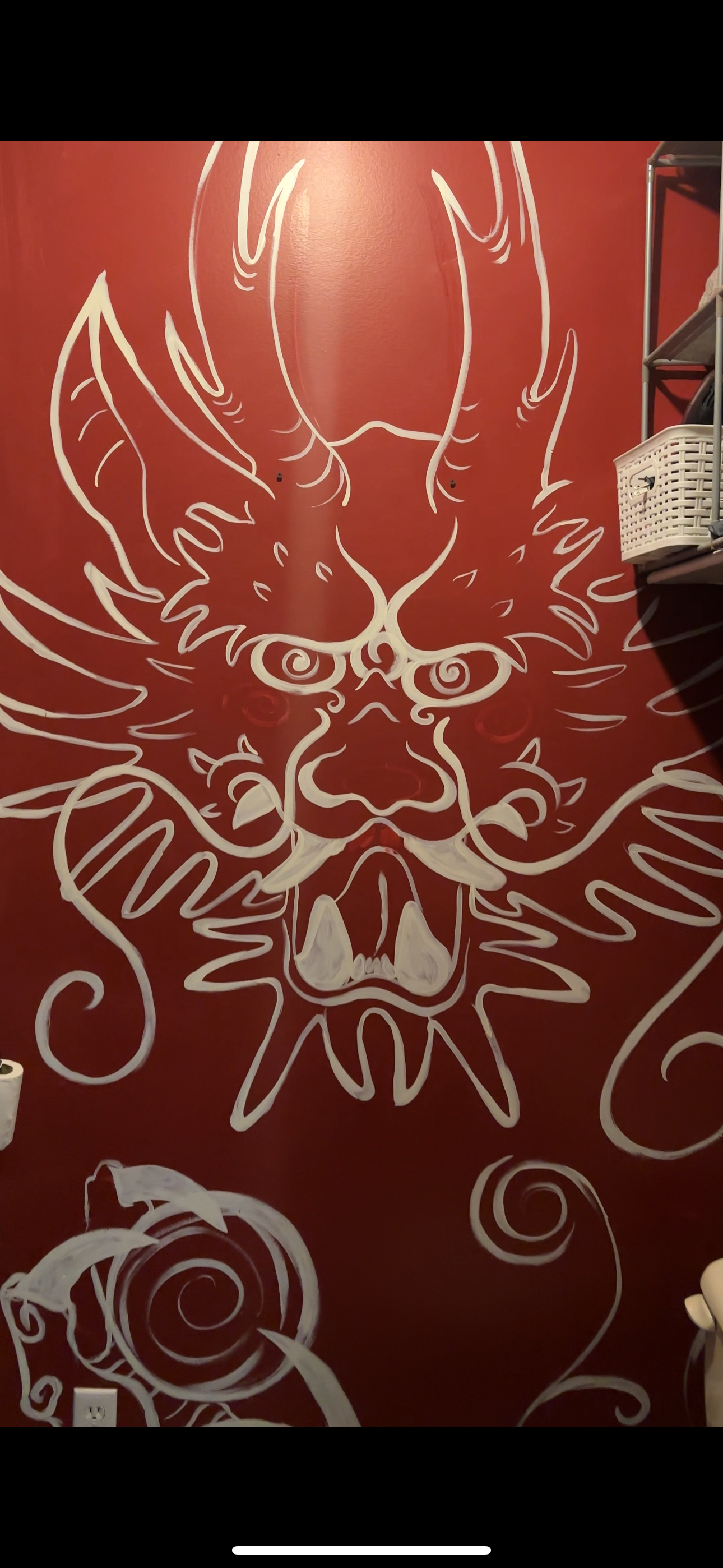 White outline of a fierce dragon or mythical creature with a large open mouth, sharp teeth, spiraled eyes, and flowing mane or flames, painted on a red wall.