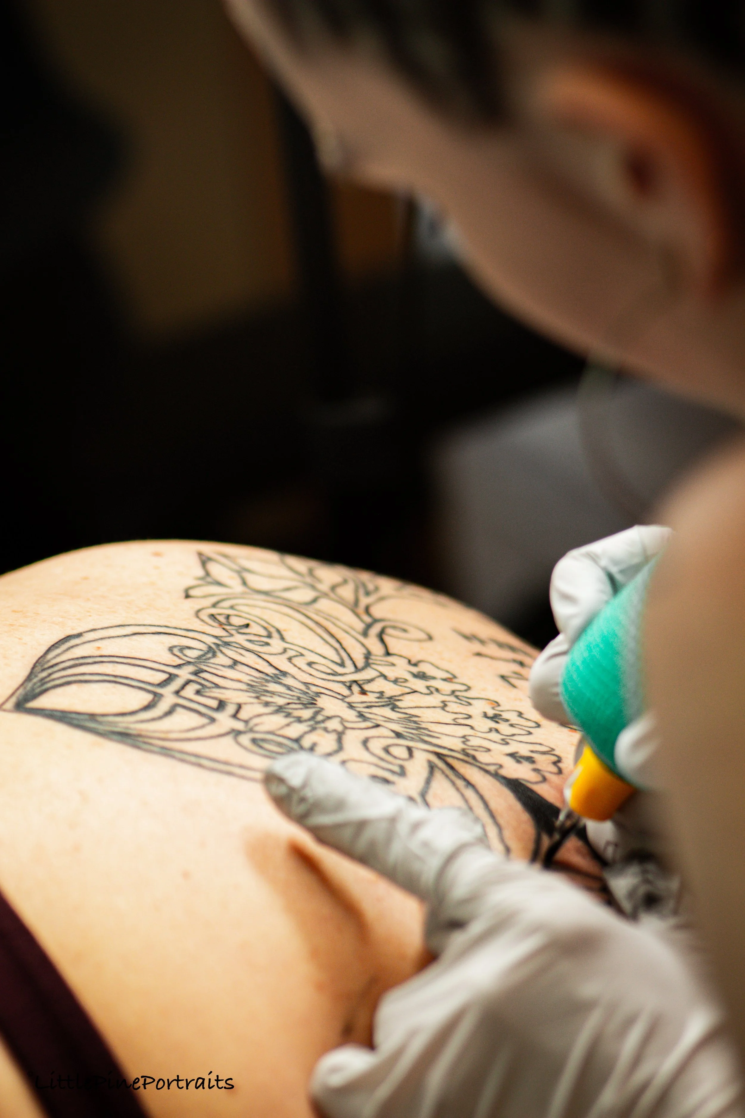 A tattoo artist is working on a tattoo design on a person's skin. The artist is wearing gloves and holding a tattoo machine. The design appears to be a detailed floral or ornamental pattern.