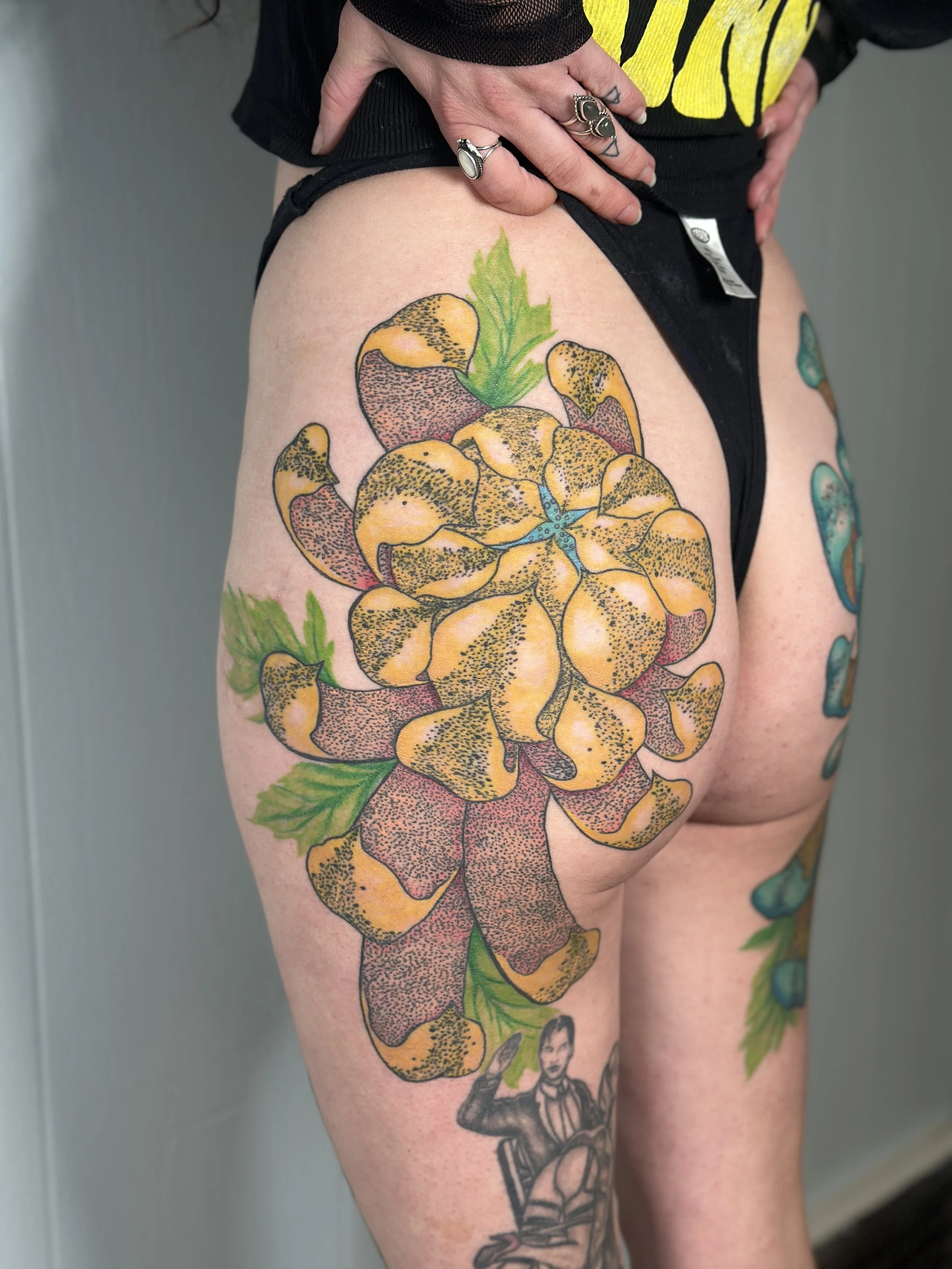 A person's upper thigh with a colorful tattoo of a detailed pineapple, yellow and brown. The person is wearing black shorts and a black shirt with yellow design, and has multiple rings and tattoos on the hand and arm.