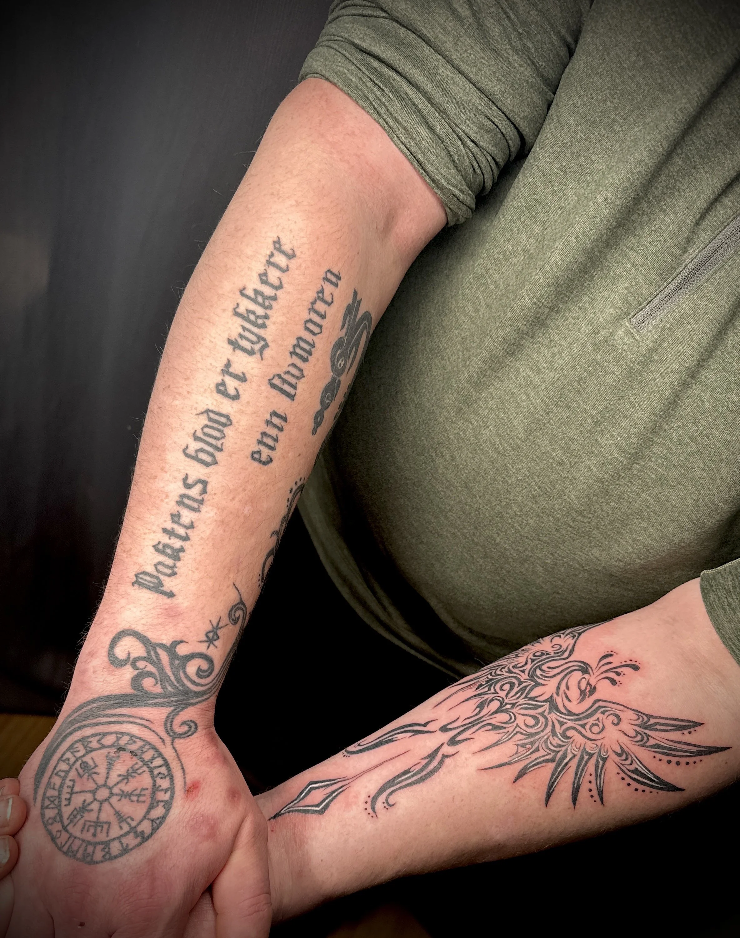 Close-up of two hands clasped together, displaying various tattoos including norse sigils, and cursive writing, with arms resting on a person's knee dressed in olive green pants.