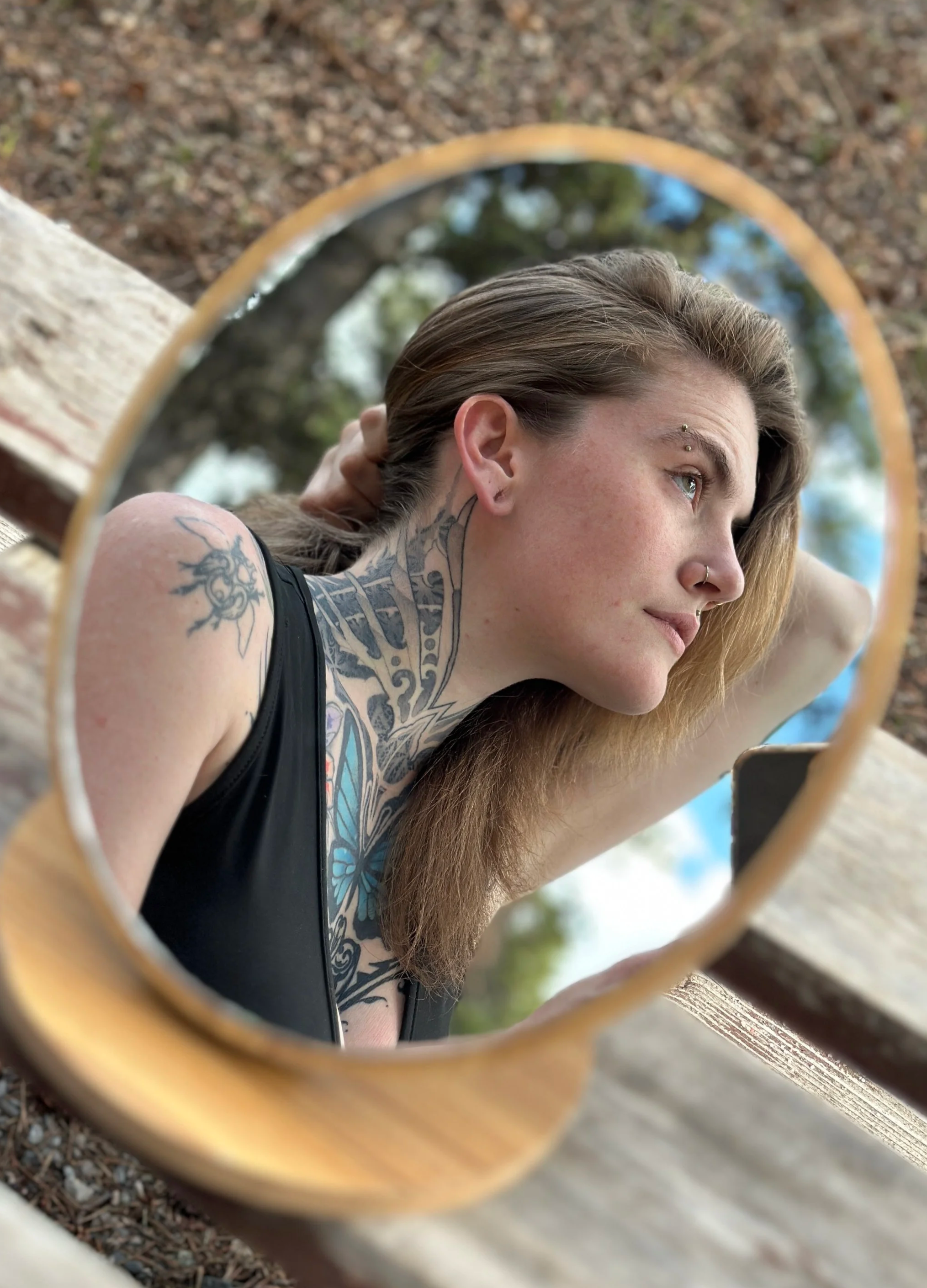 A young woman with a tattoo on her shoulder and a nose ring is seen through a round mirror reflecting her face and part of her upper body. She appears to be outdoors with trees and sky visible in the background.