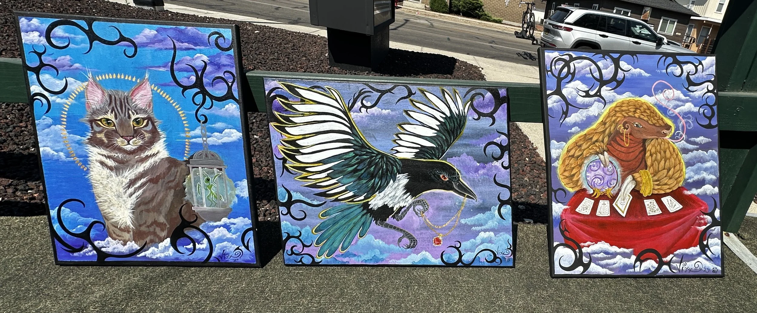 Three colorful paintings displayed outdoors, featuring a cat with a halo and lantern, a bird with jewelry flying against a cloudy sky, and an anthropomorphic lion dressed as a fortune-teller surrounded by tarot cards.