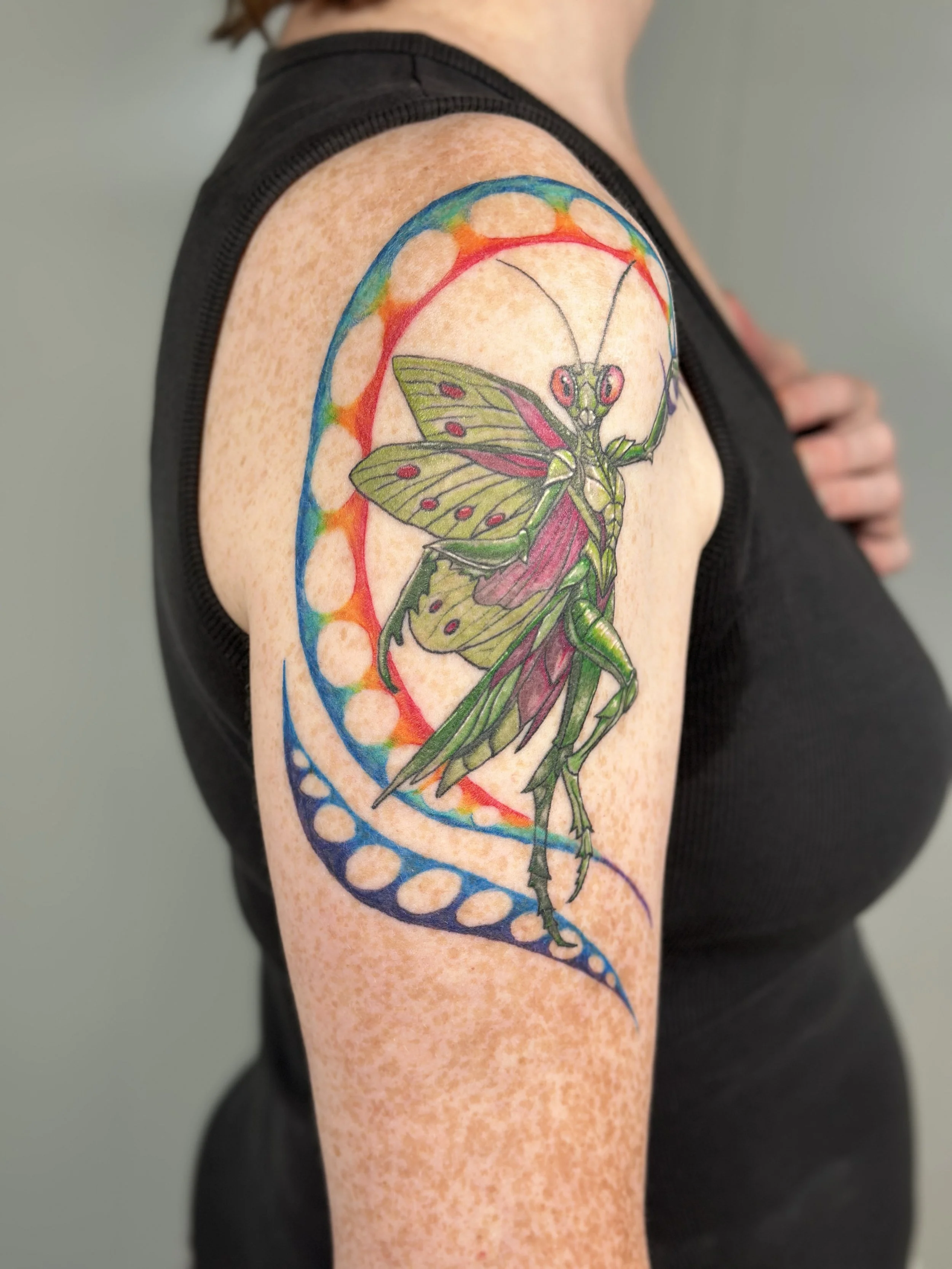 Colorful tattoo of a praying mantis on a person's upper arm, featuring geometric shapes and rainbow-like colors.