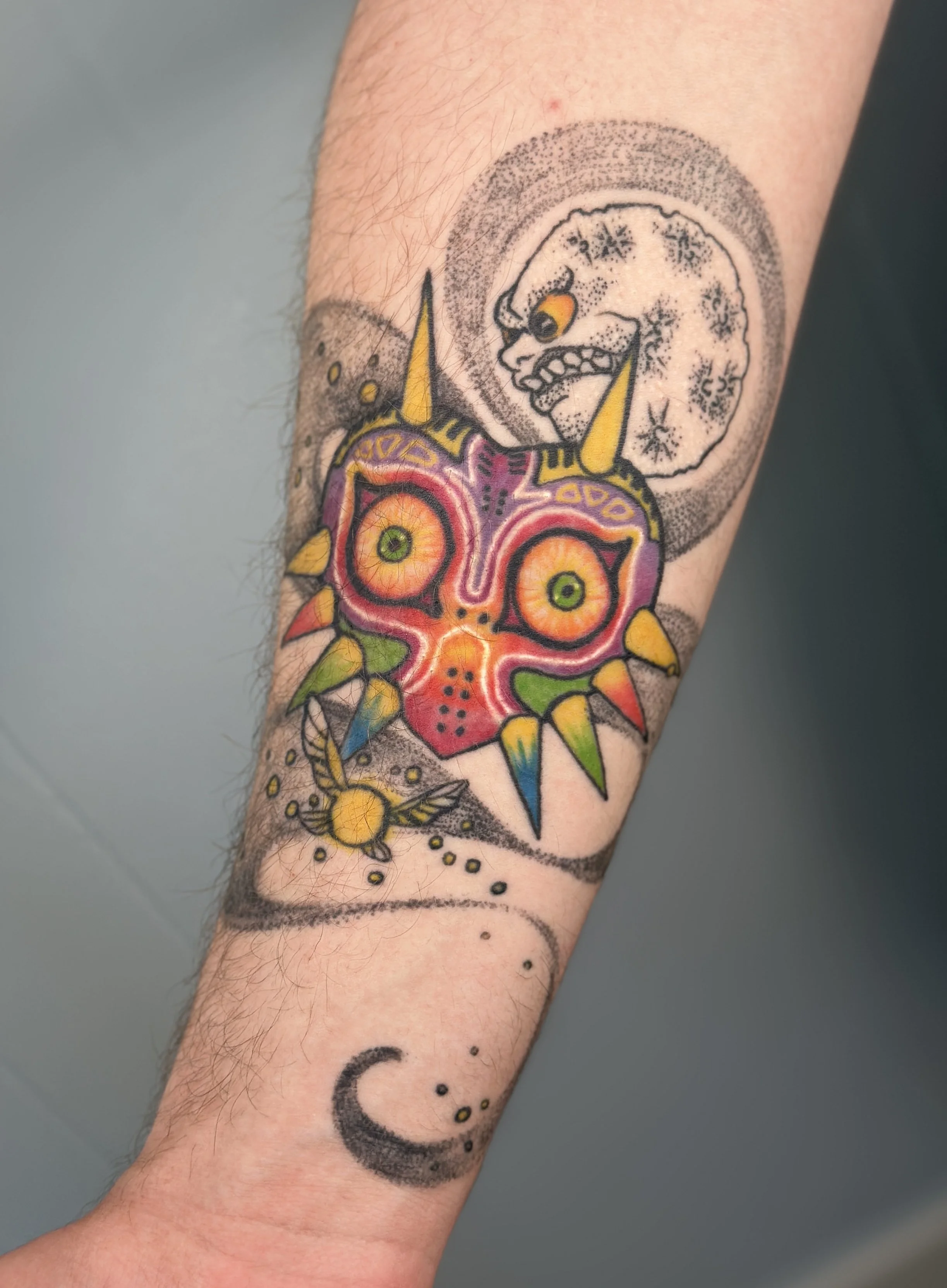 Colorful tattoo on arm featuring a creature with a pink face, large yellow horns, and eyes with green and orange irises, against a background of gray ink dots and lines.