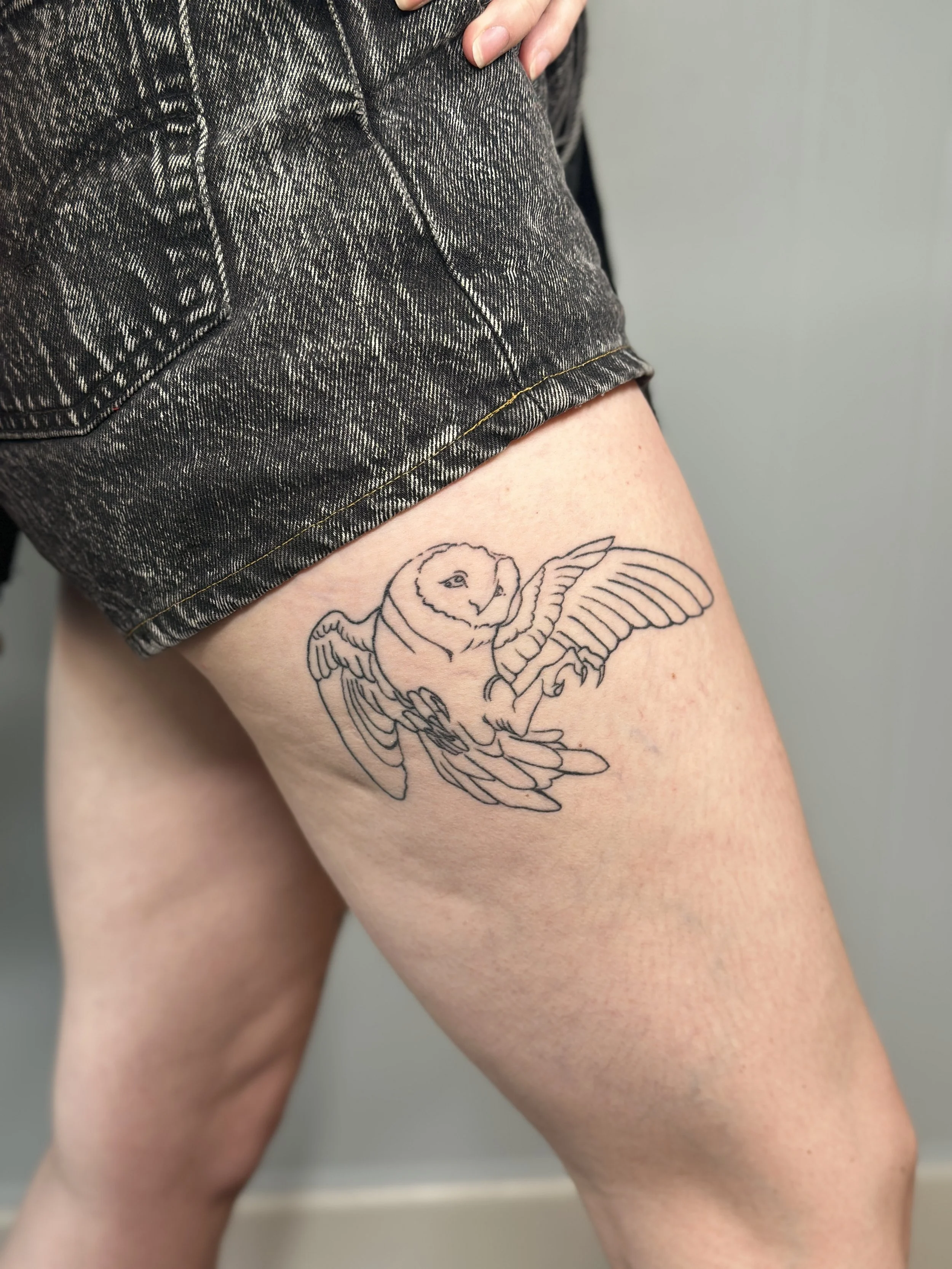 A person wearing black denim shorts with a tattoo of an owl with outstretched wings on their upper thigh.