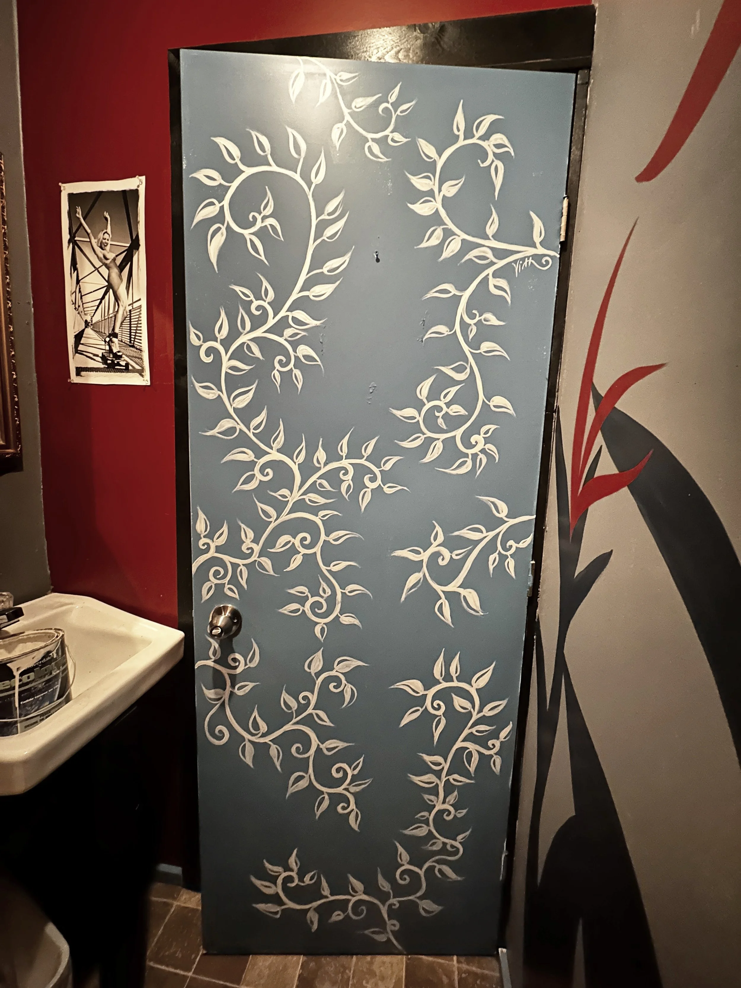 A restroom stall door painted light blue with white swirling vine and leaf design. Next to the door is a small white sink and a poster of a woman skateboarding on a bridge.