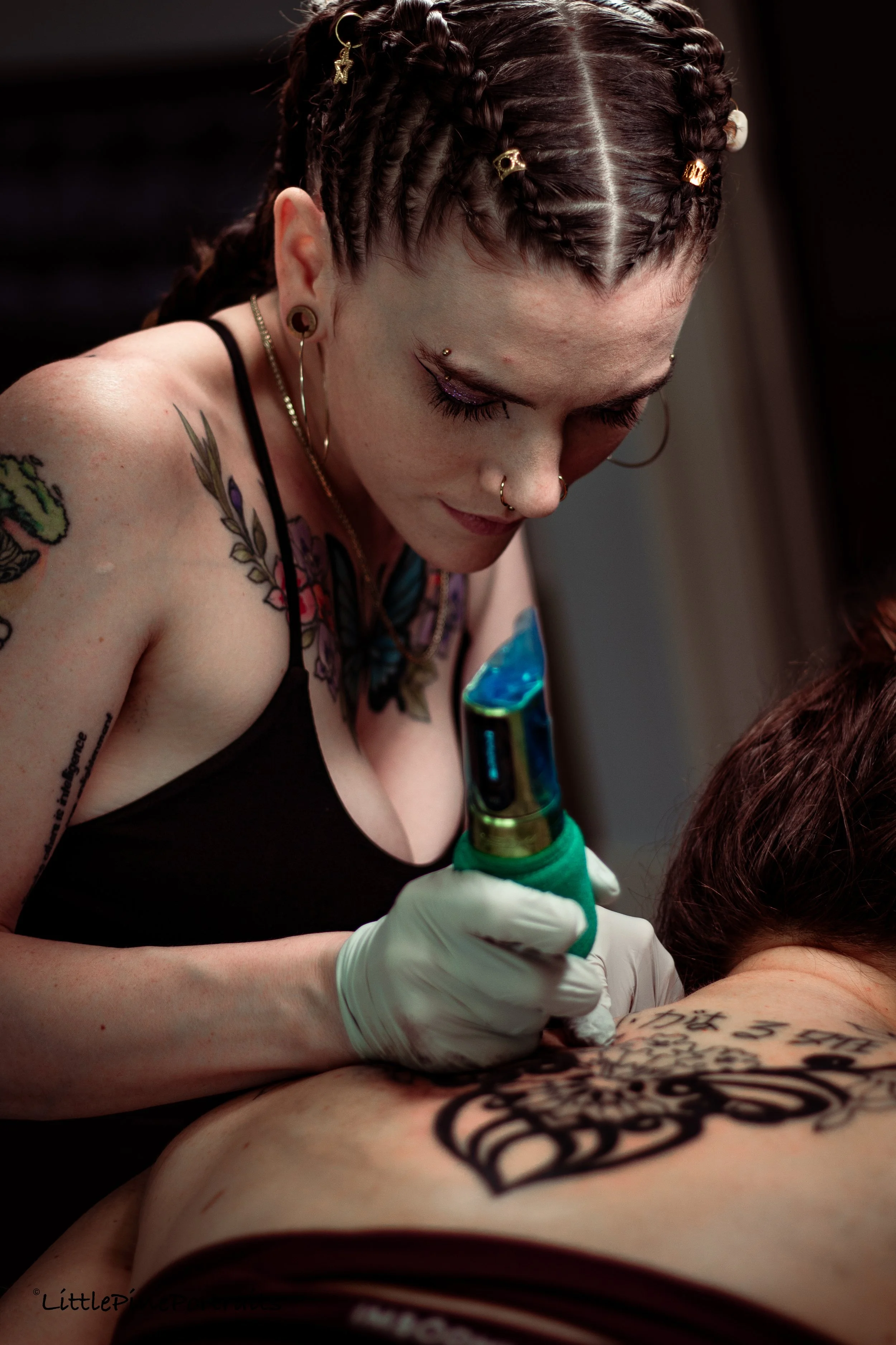 A woman with braided hair, tattoos, and piercings is tattooing a body with a tattoo machine, wearing gloves, and focused on her work.
