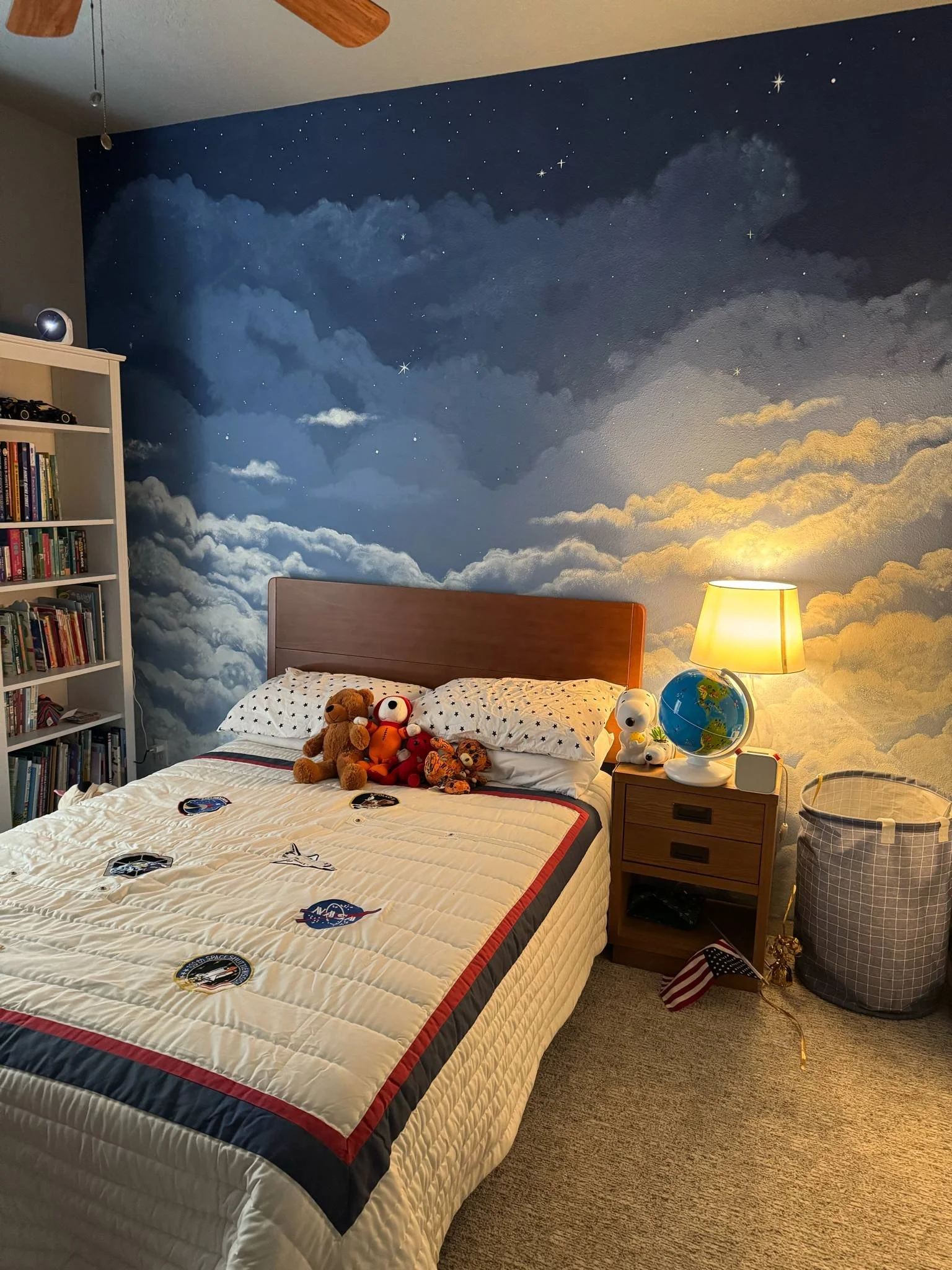 A bedroom with a space-themed cloud mural on the wall behind the bed, a white bookshelf filled with books and toy cars, a nightstand with a globe, a lamp, and stuffed animals, a laundry basket, and a ceiling fan.