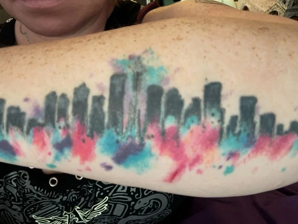Colorful watercolor tattoo of a city skyline on person's upper arm.