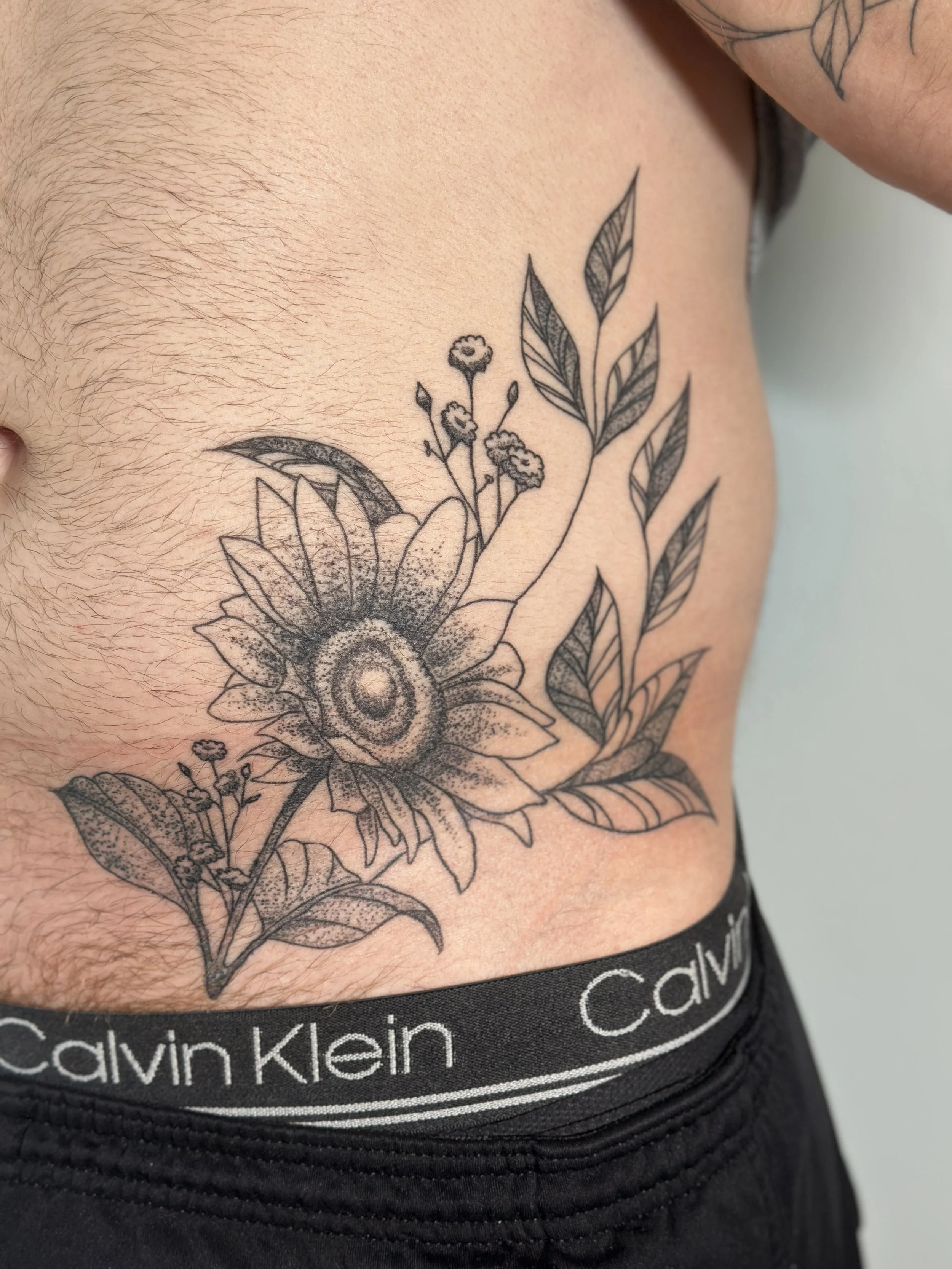 Close-up of a person's side showing a black and gray floral tattoo and wearing black Calvin Klein underwear.