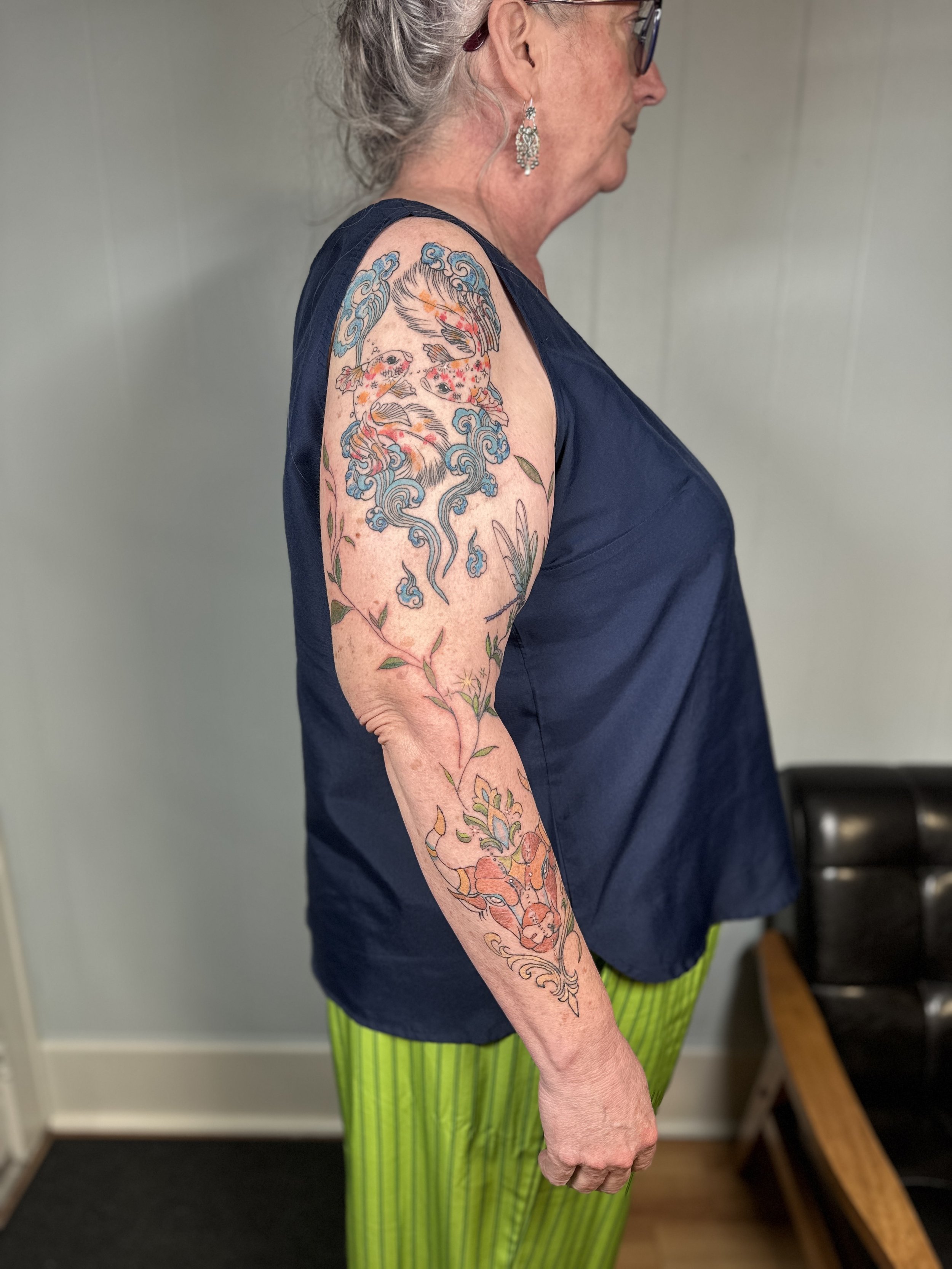 A side view of an elderly woman with gray hair tied back, wearing glasses, earrings, a dark sleeveless top, and bright yellow-green striped pants. She has a large colorful tattoo of koi fish, waves, and floral designs on her upper arm.