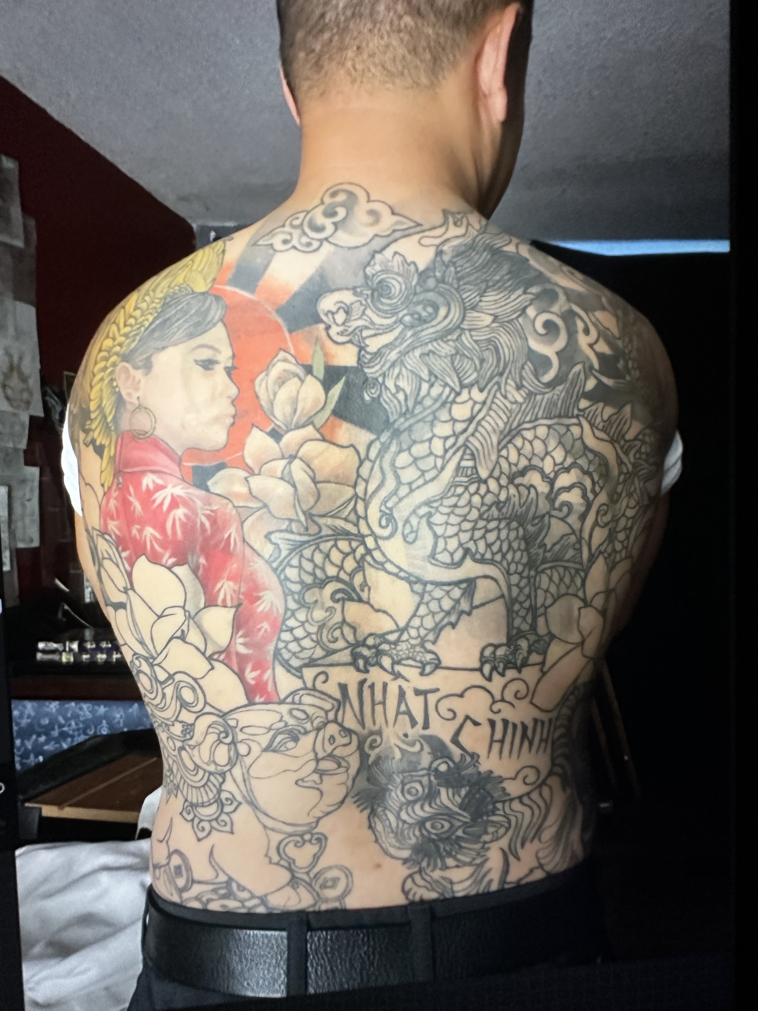 Back view of a person with a large, detailed tattoo covering their back and shoulder. The tattoo features a woman with a braid, a dragon, a tiger, and various floral and cloud elements. The woman is wearing a red garment with white floral patterns. There is also text in the tattoo, but part of it is obscured.