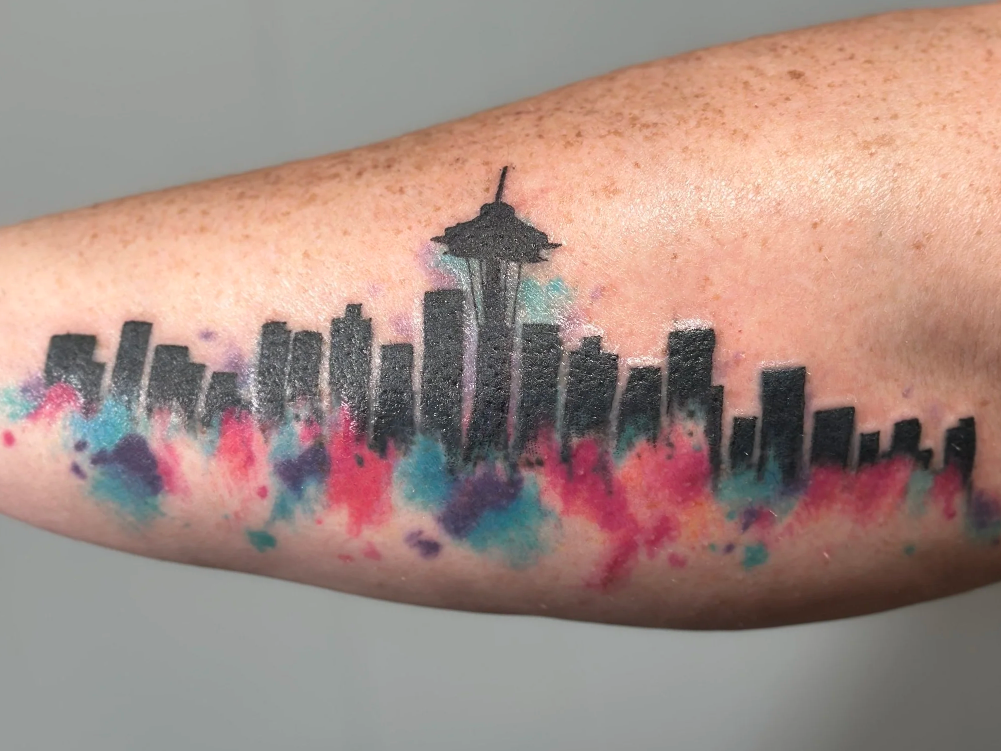 Close-up of a tattoo featuring the Seattle Space Needle with a city skyline silhouette beneath it, surrounded by colorful watercolor-style splashes.
